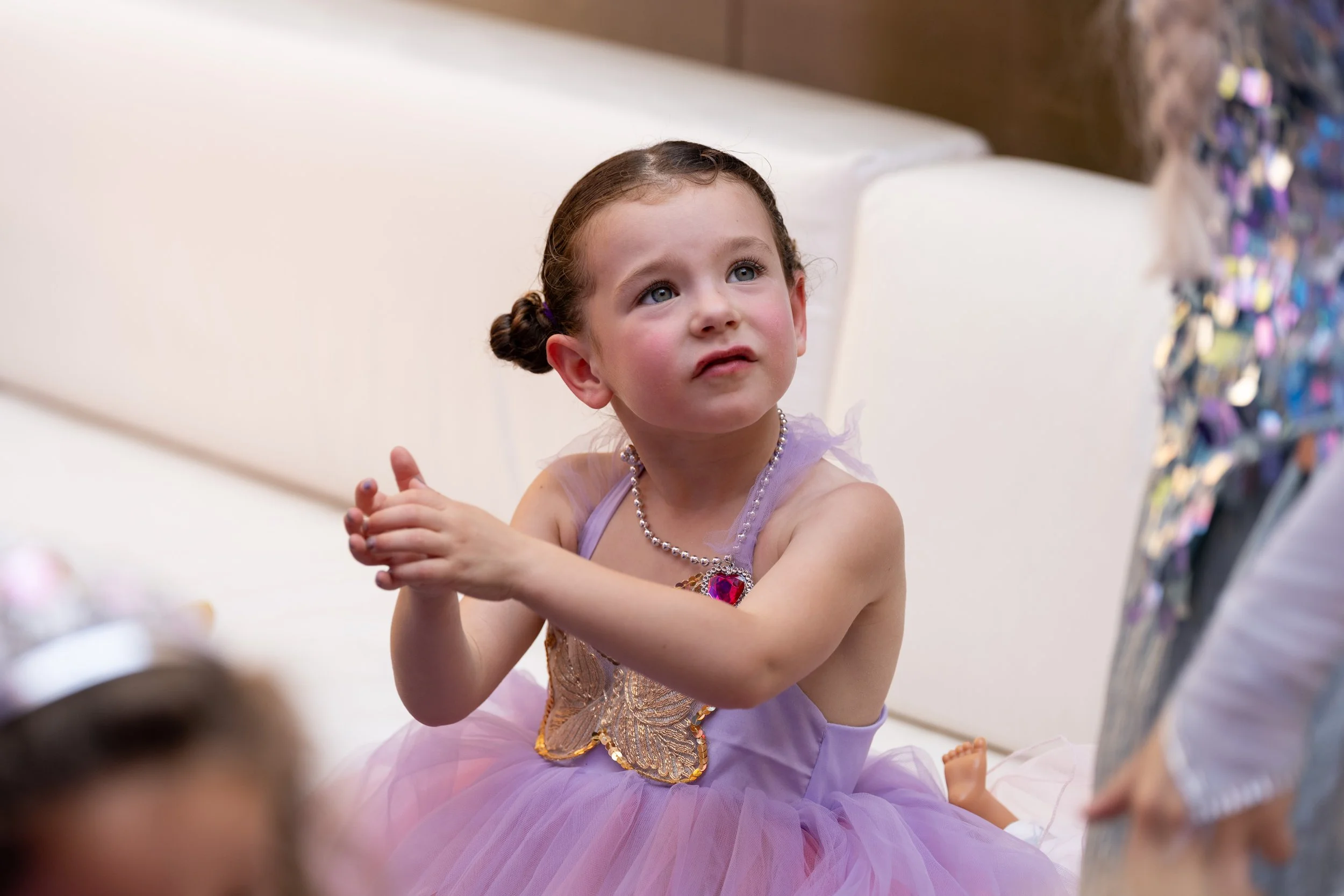 A young girl in a purple tutu dress with butterfly embroidery and a beaded necklace sitting on a white sofa, looking up with a curious and slightly puzzled expression, during a festive event.