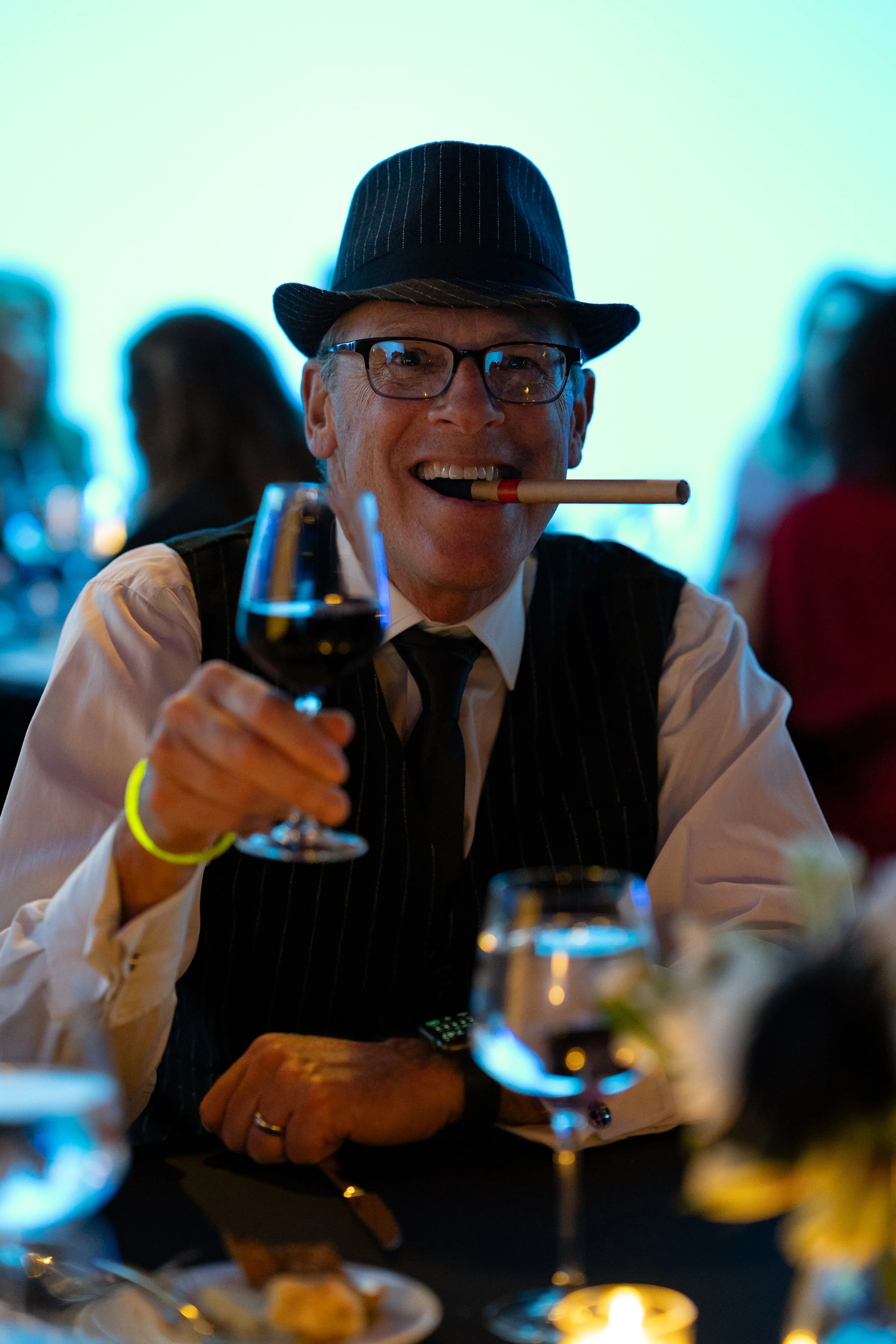 A man wearing a hat, glasses, and a vest, smiling with a cigar in his mouth, holding a glass of red wine at a social event.