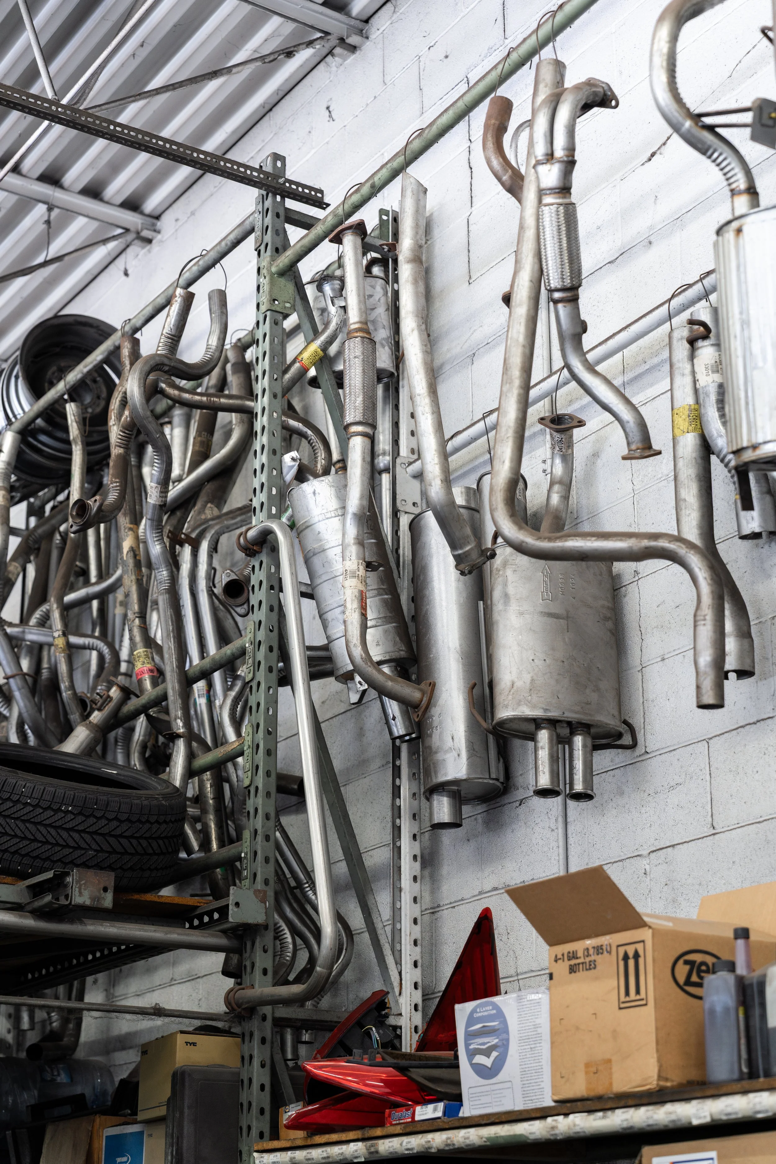 A wall in a workshop with various used metal car exhaust pipes and mufflers hanging on hooks.