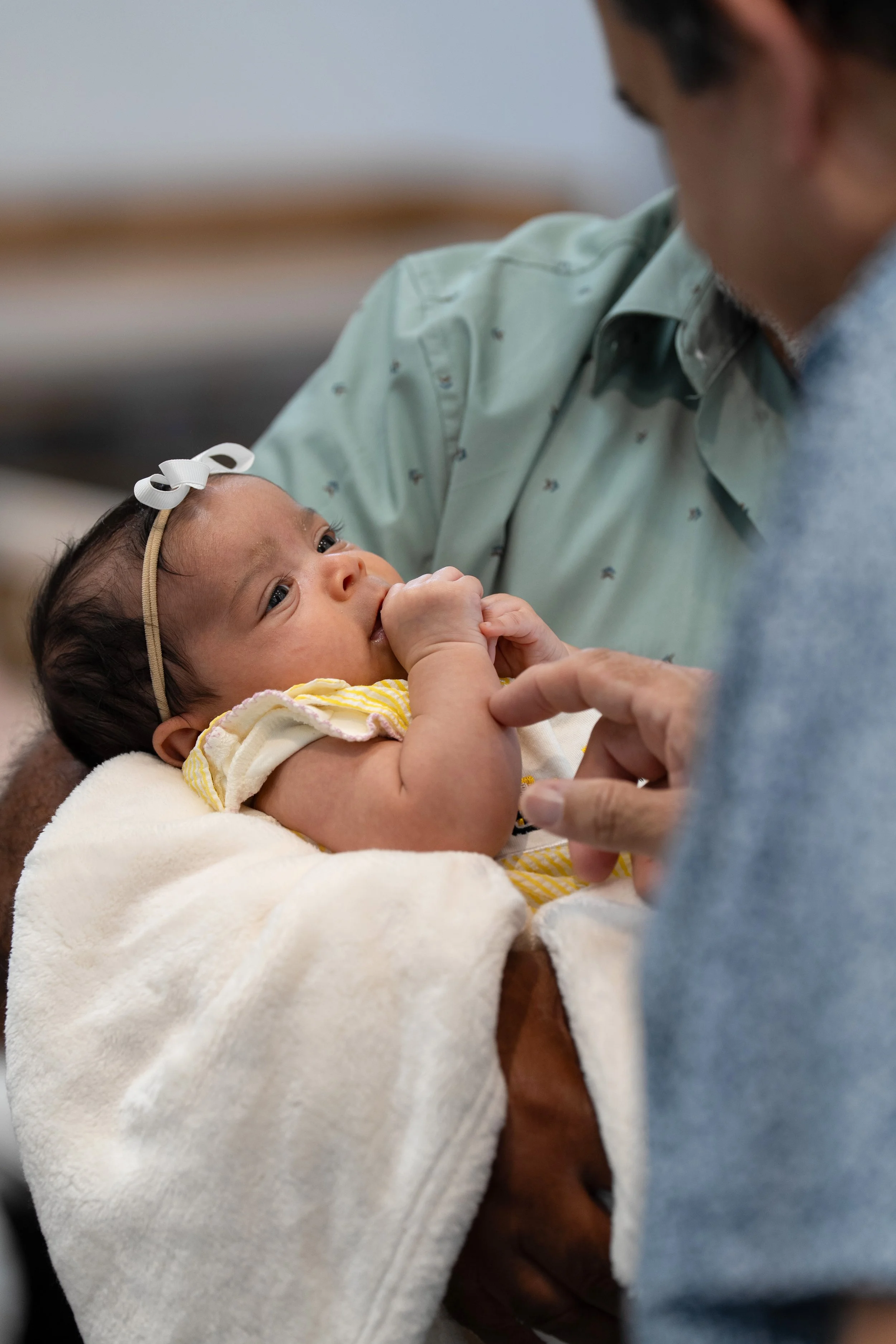 A baby lying on a person's arm, with someone else gently holding the baby's hand. The baby is sucking on its thumb and has a white bow in her hair.