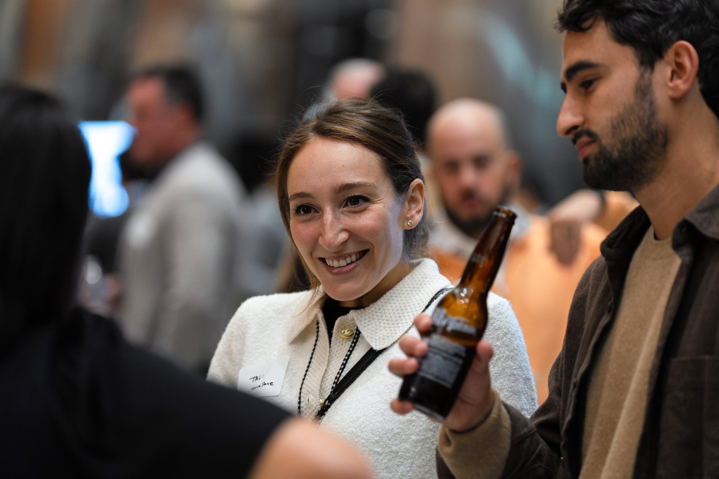 A woman smiling and talking to a man holding a beer bottle at a social gathering or party.