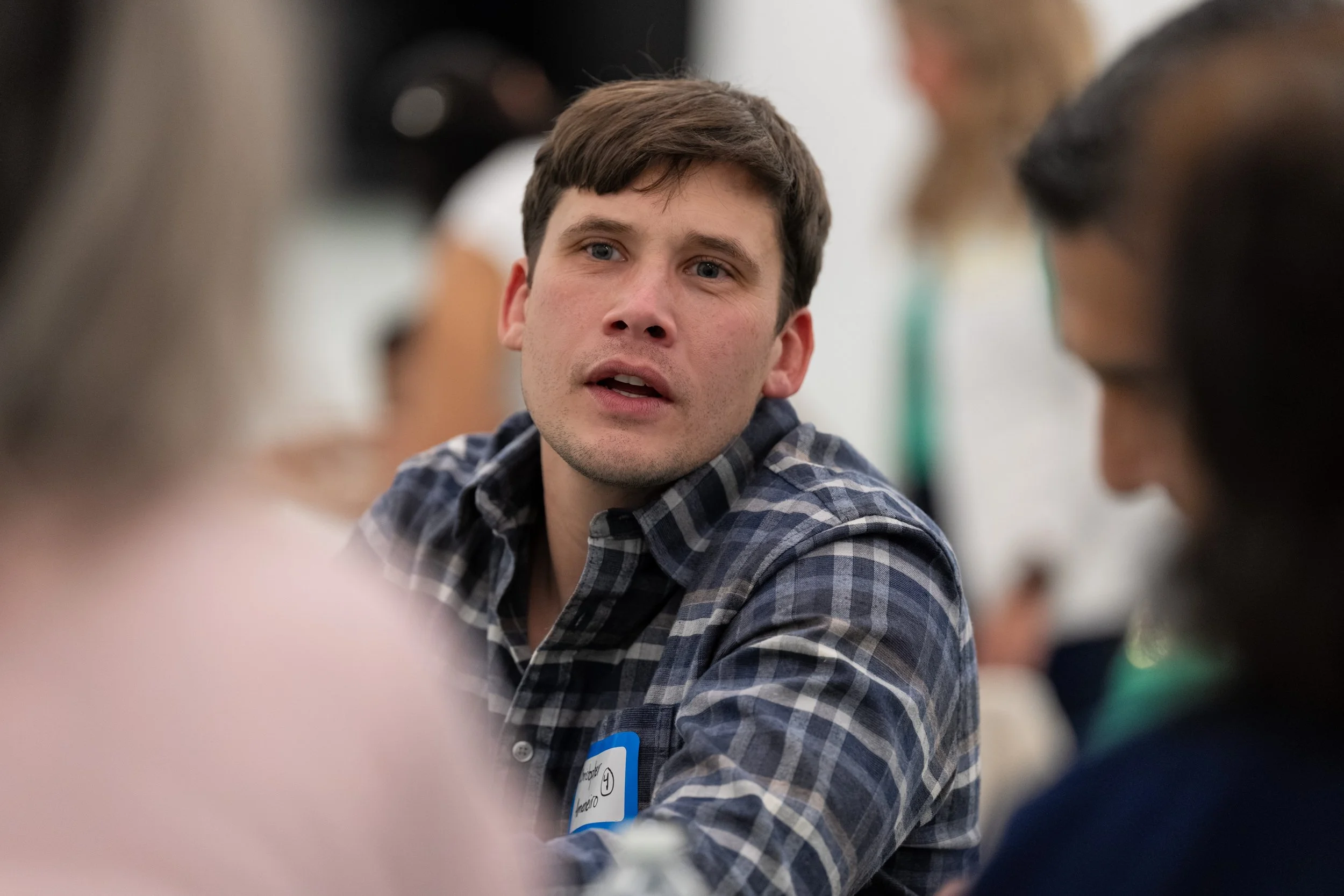 Young man in a plaid shirt speaking and listening at a social gathering or workshop, with others around him.