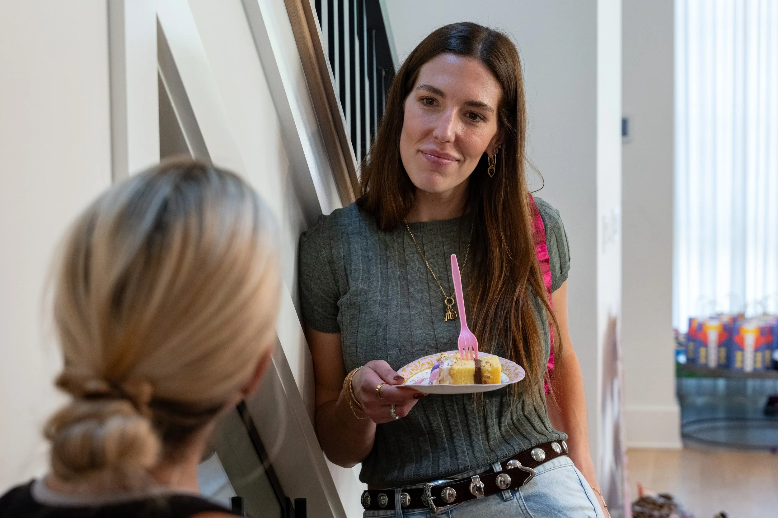 A woman with long brown hair, wearing a green top and jewelry, holding a plate with a slice of cake and a pink fork, standing indoors and talking to a woman with blonde hair tied back.