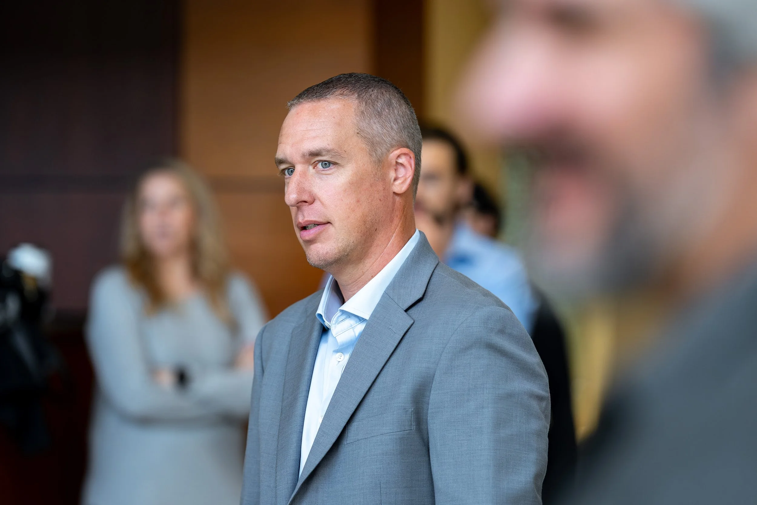 A man with short hair and a serious expression wearing a gray suit and light blue shirt, standing at a professional event with blurred people in the background.
