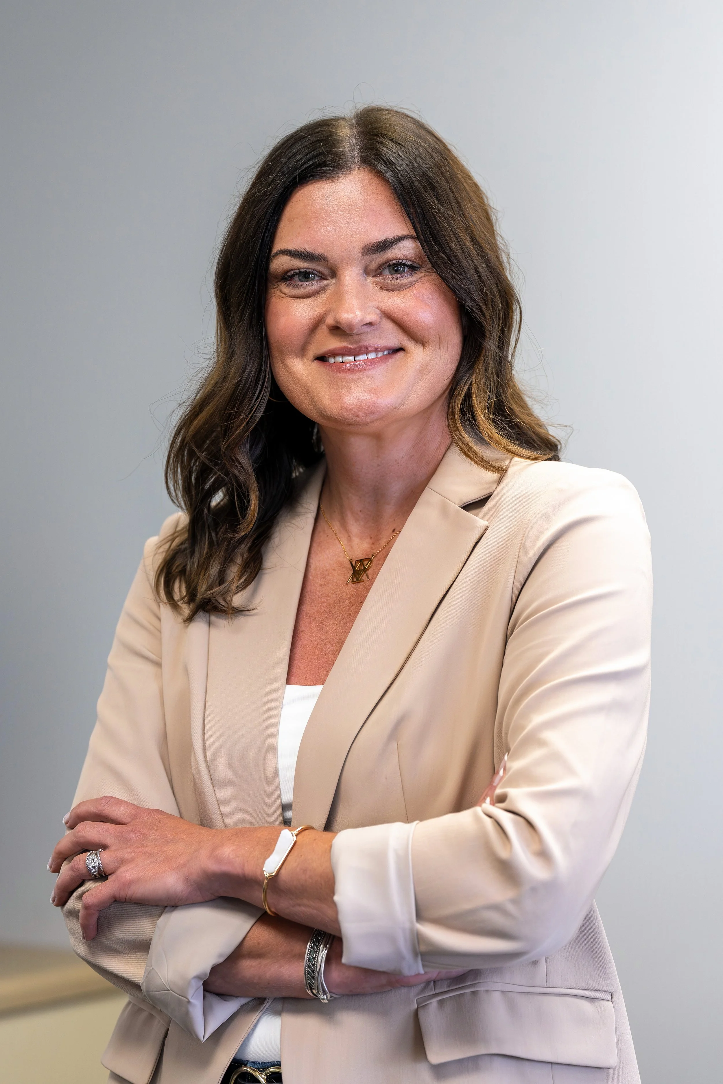 A woman with shoulder-length dark hair, wearing a beige business suit, smiling with arms crossed, standing against a neutral background.