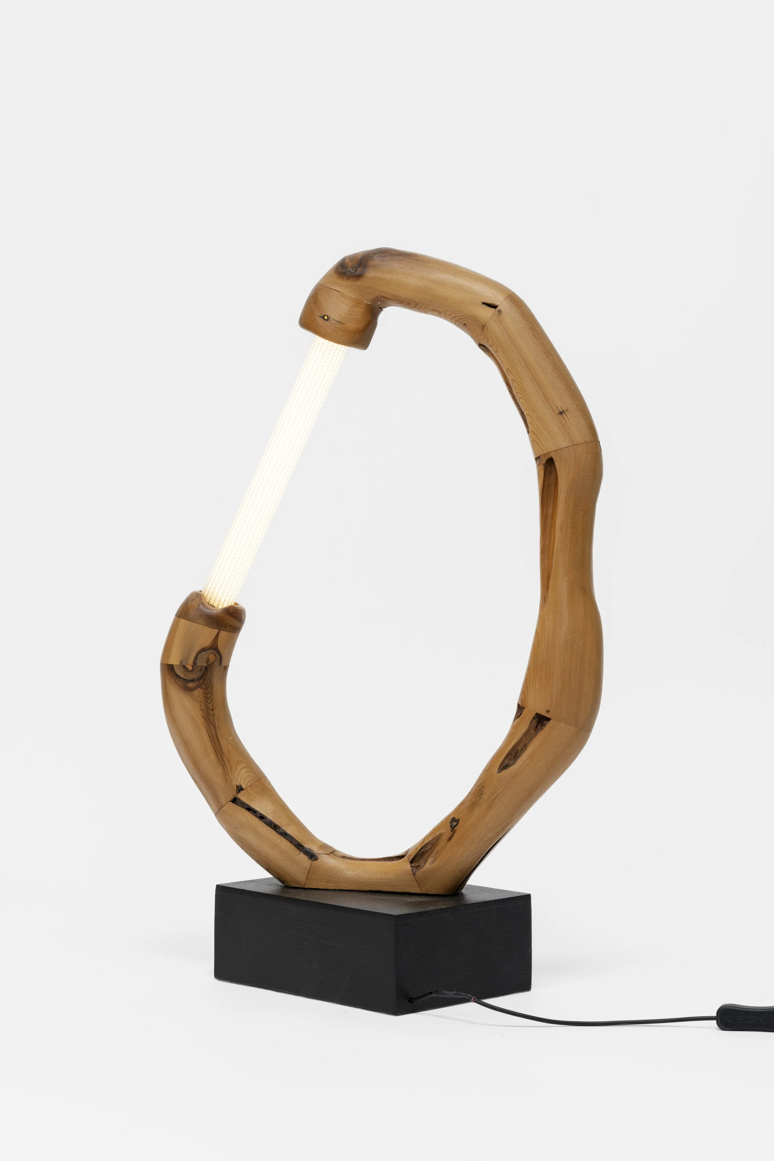 A lamp with a frame made of curved, natural wood and a black base, featuring a white, textured light strip as the light source, connected by an electrical cord.