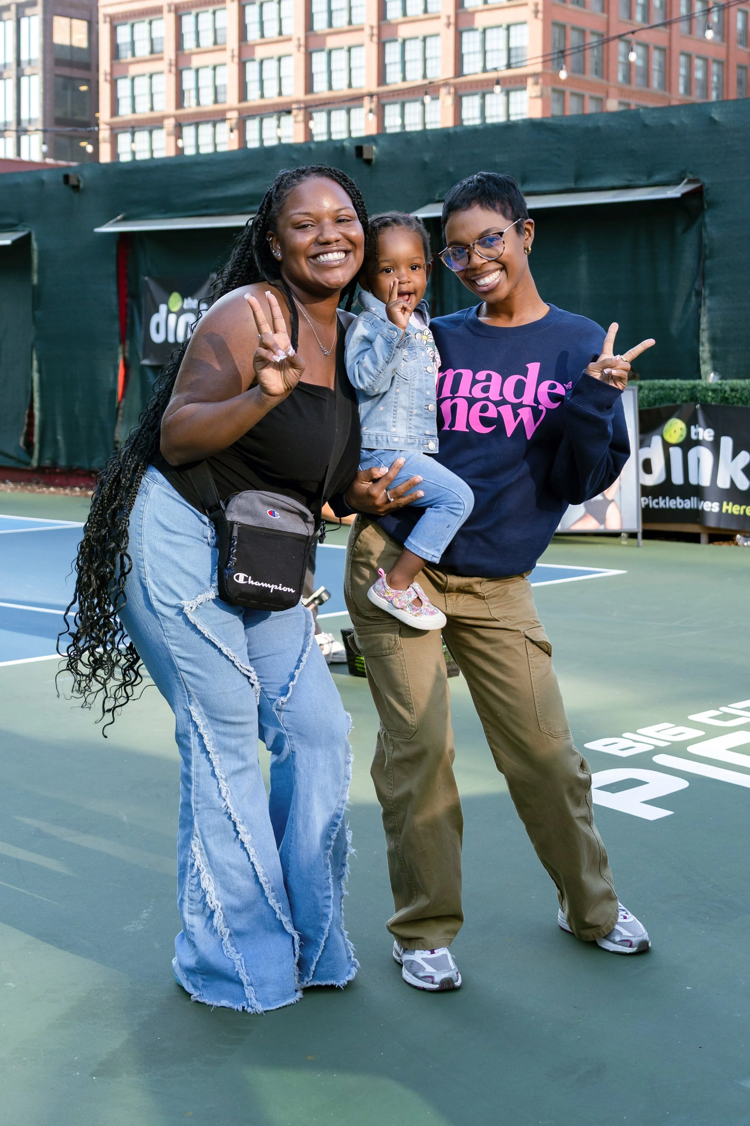 Three women and a young girl smiling and making peace signs at an outdoor tennis court, with a banner in the background that reads 'the dink' and 'Pickleballers Here'.