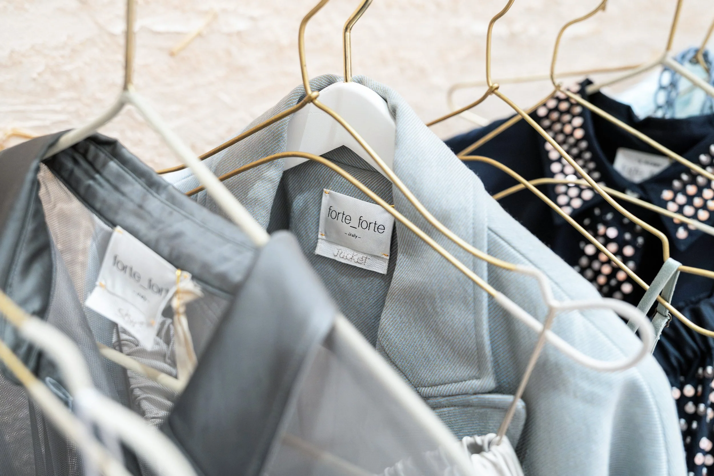Clothing rack with gray and navy jackets on gold hangers, featuring brand tags 'forte forte' and 'loree lory'