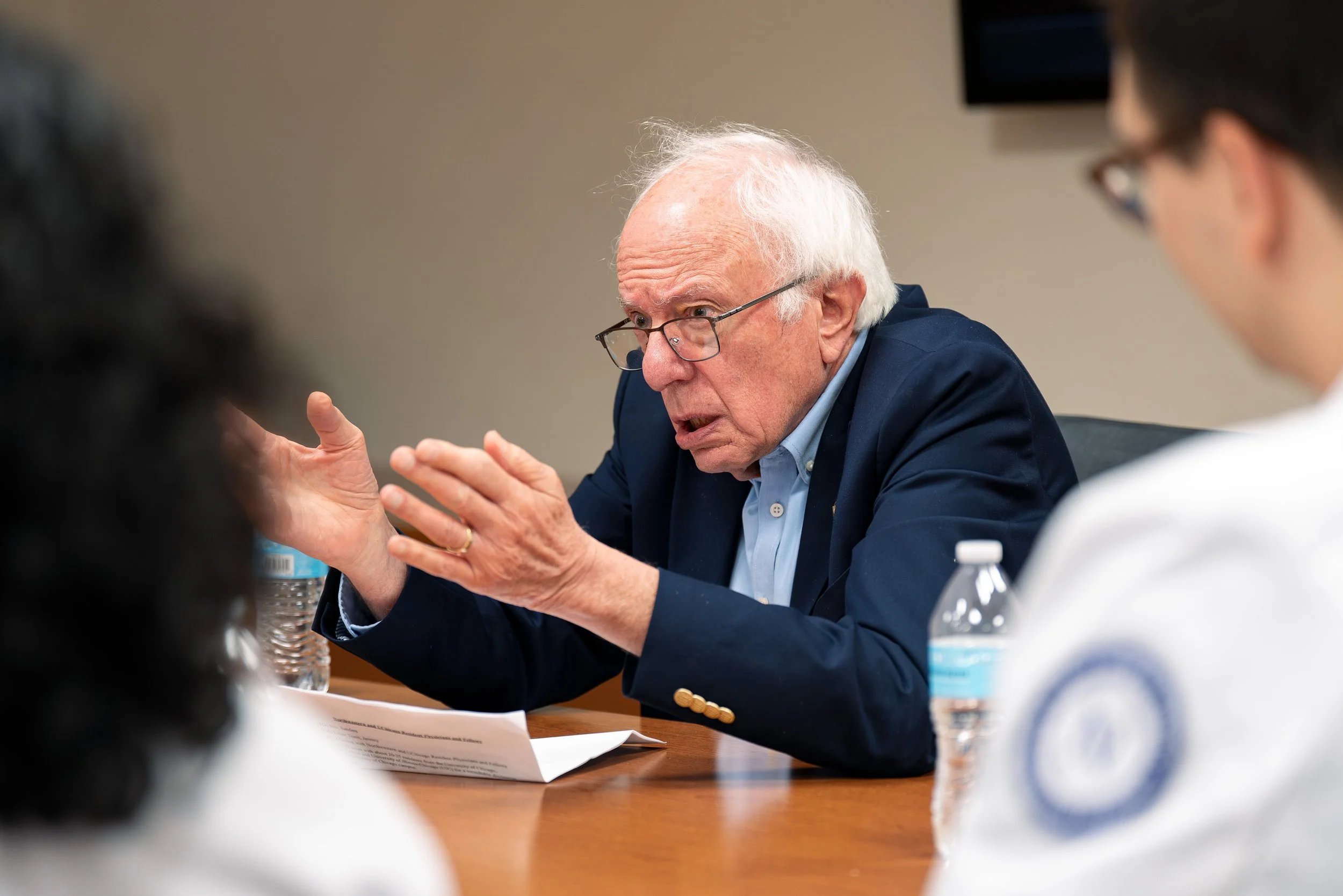 An elderly man with white hair, glasses, and a dark blue blazer is speaking and gesturing with his hands during a meeting or discussion, with a water bottle and papers in front of him. Two people, one partially visible on the left and another on the 