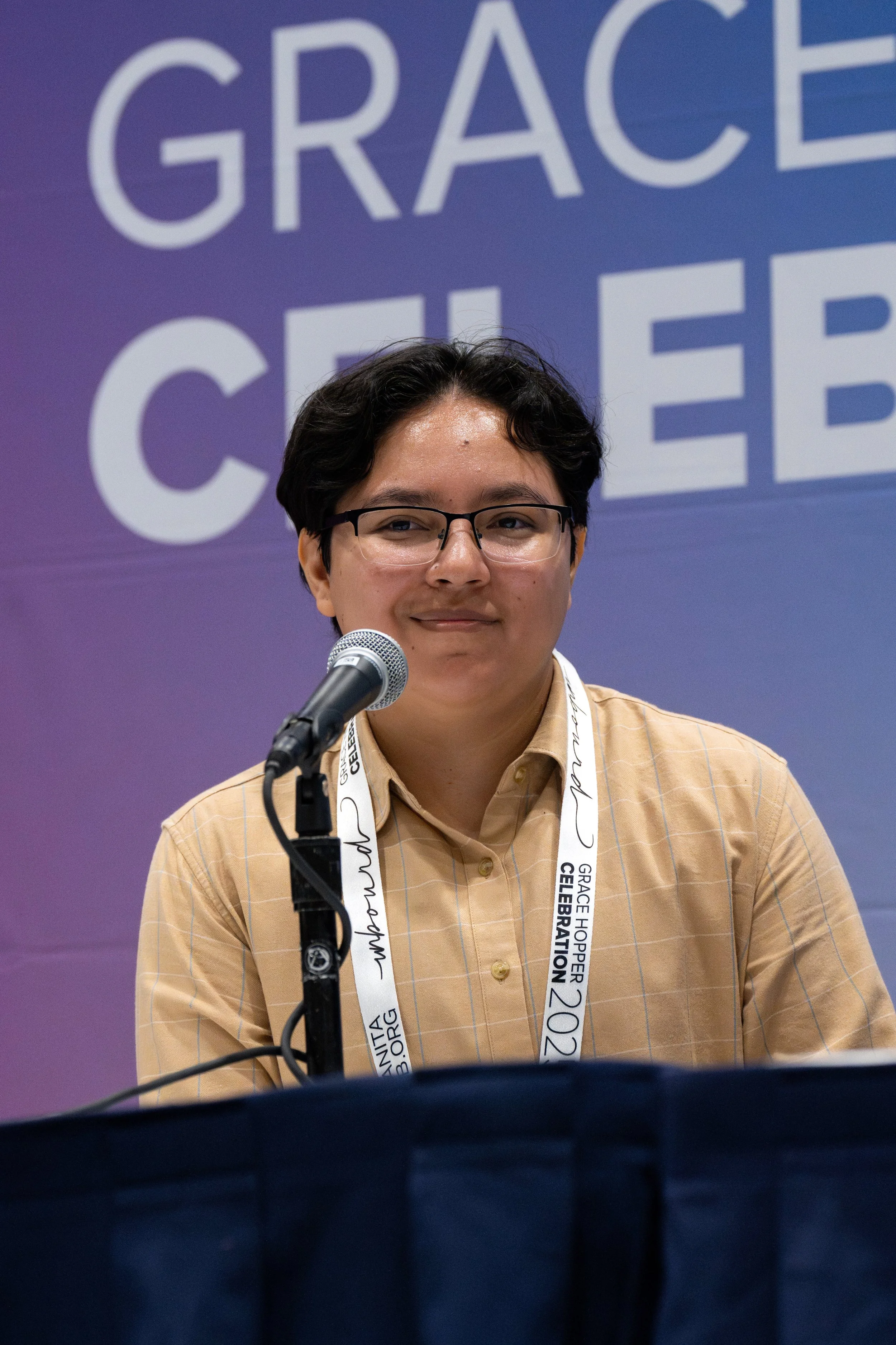 A person with short dark hair and glasses is smiling and sitting behind a microphone at a conference or event. They are wearing a beige shirt and a lanyard with text that reads 'Grace Hopper Celebration 2023'.