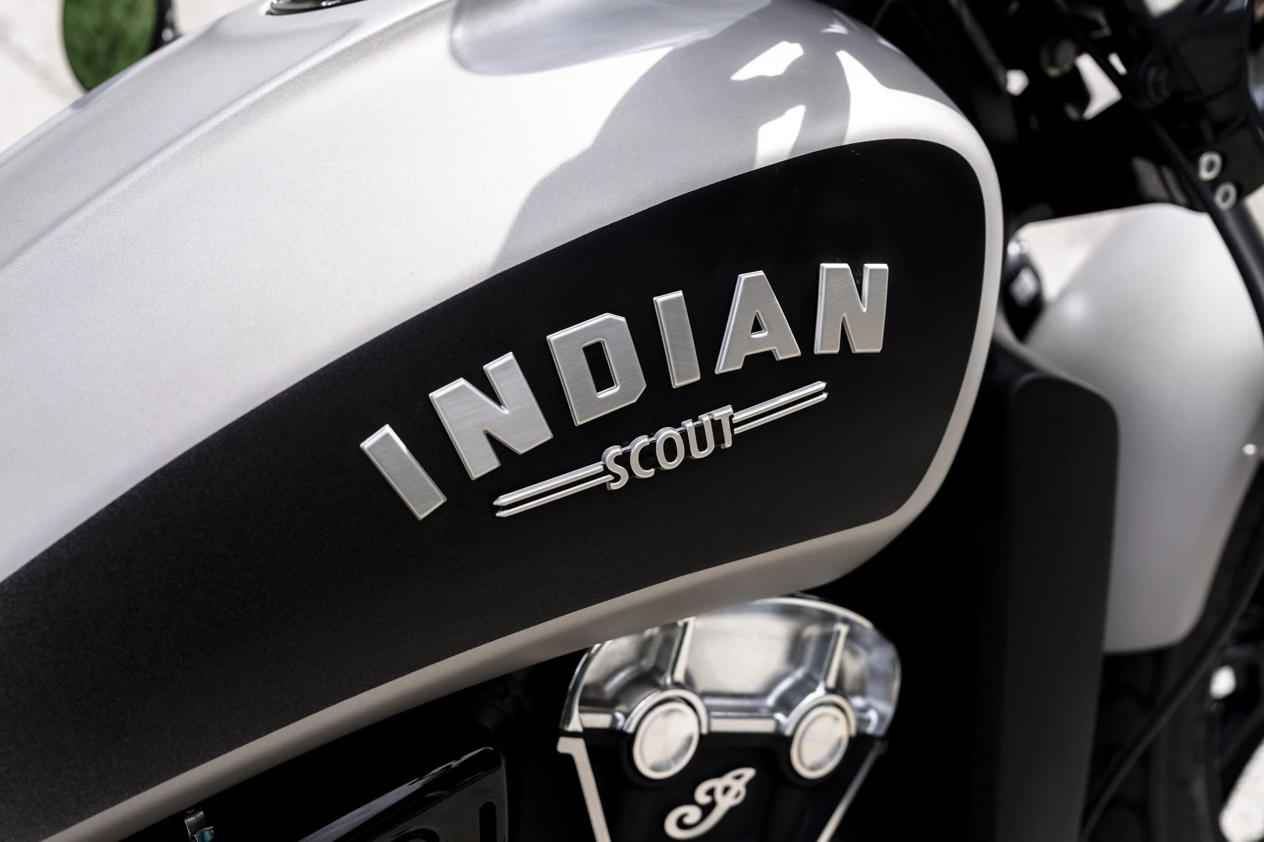 Close-up of an Indian Scout motorcycle, focusing on the black and silver branding on the fuel tank, with parts of the engine and frame visible.