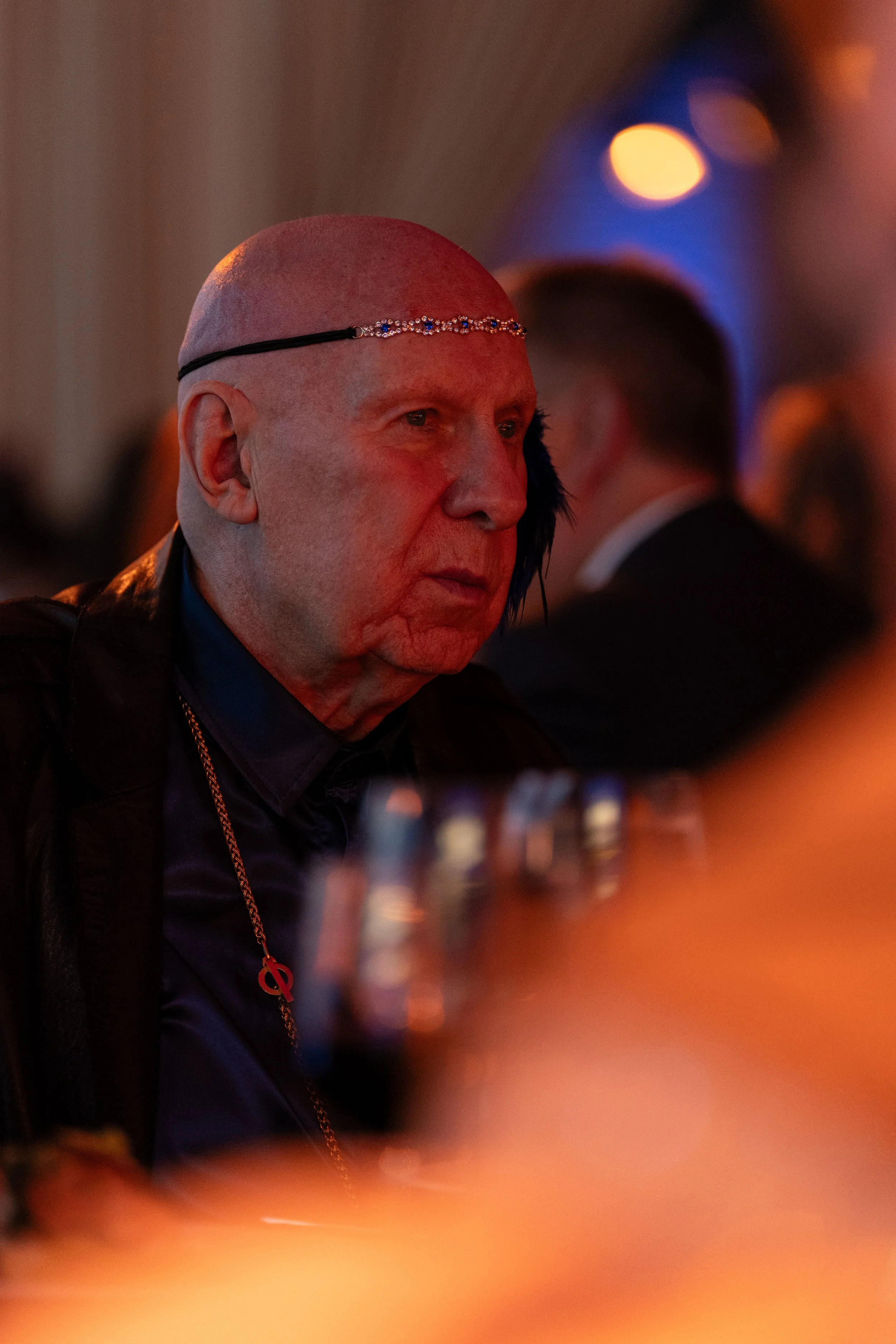 A man with a bald head, wearing a headband with decorative jewels, is seated at a table with glasses, in a dimly lit environment, possibly a social event or dinner party.