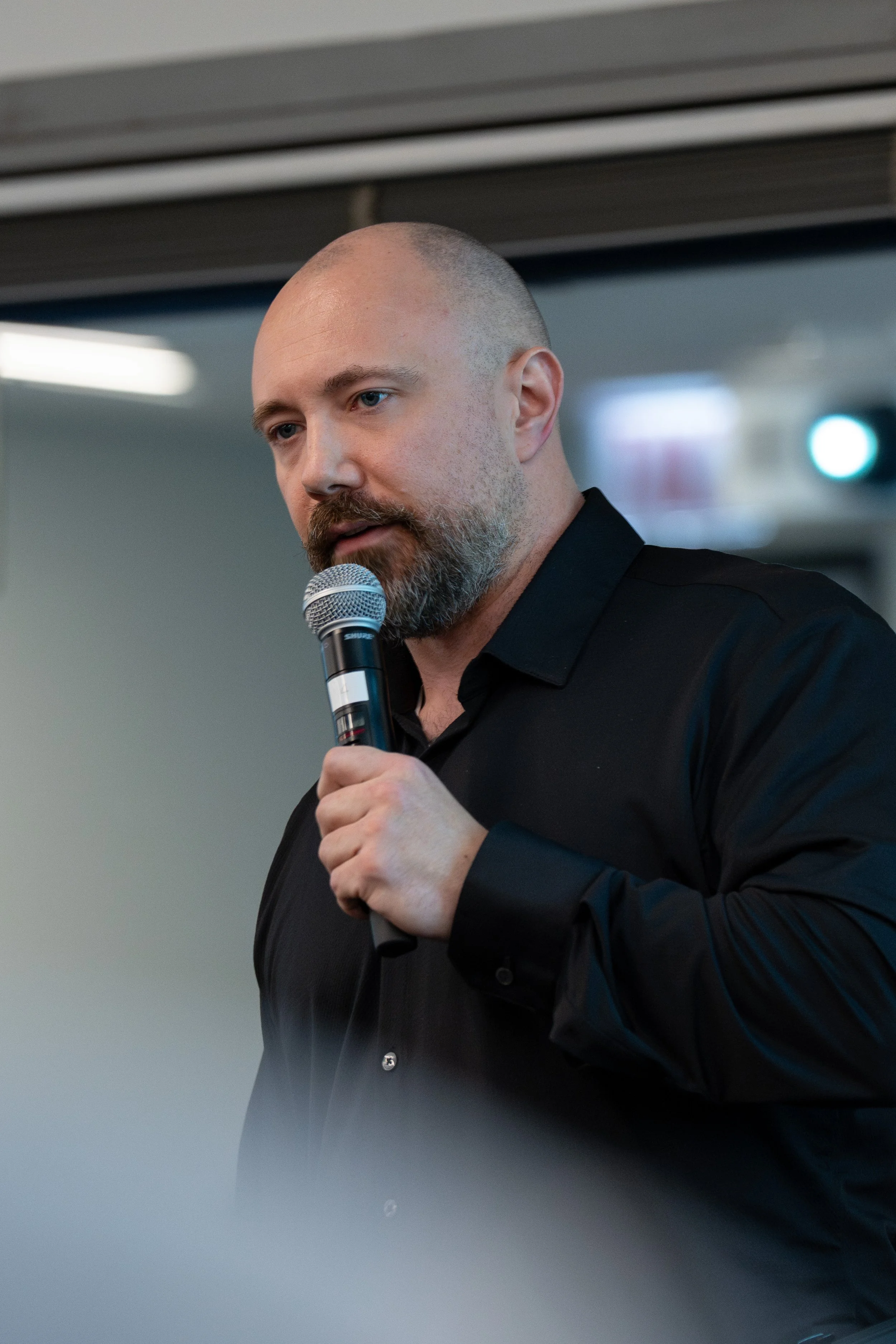 A man with a beard and shaved head, wearing a black shirt, holding a microphone and speaking.