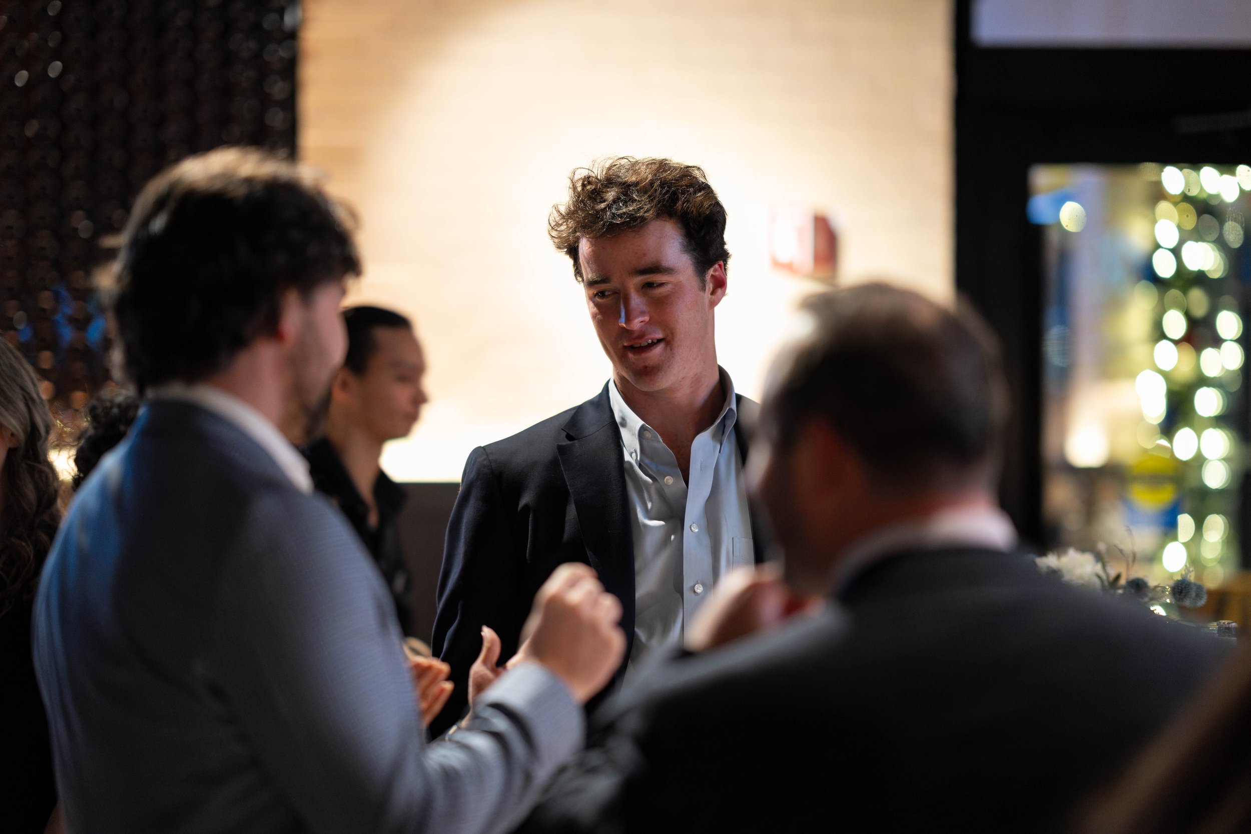 A group of people dressed in formal attire engaged in conversation at an indoor event with warm lighting and a blurred background.