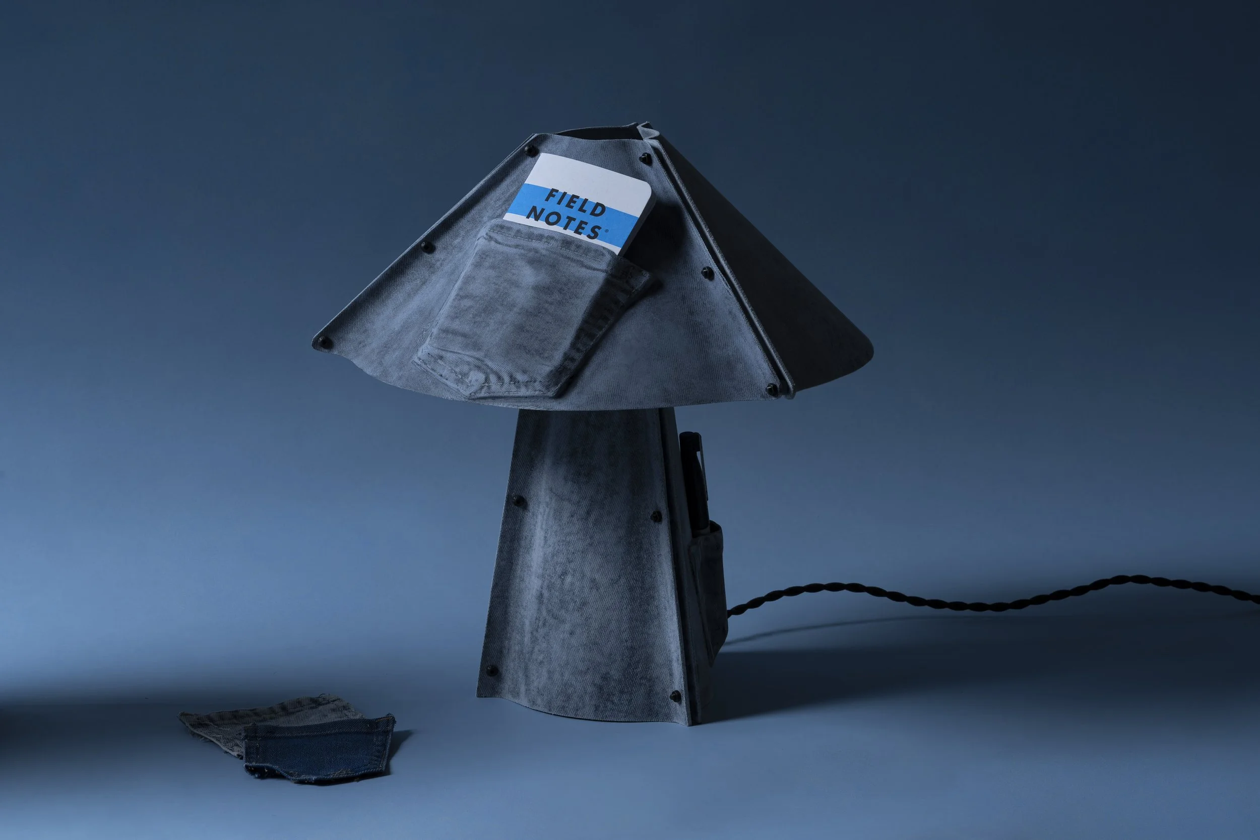 Upward view of a metallic streetlamp with a 'Field Notes' notebook tucked in a pocket on its side, against a dark blue sky with a layer of clouds.