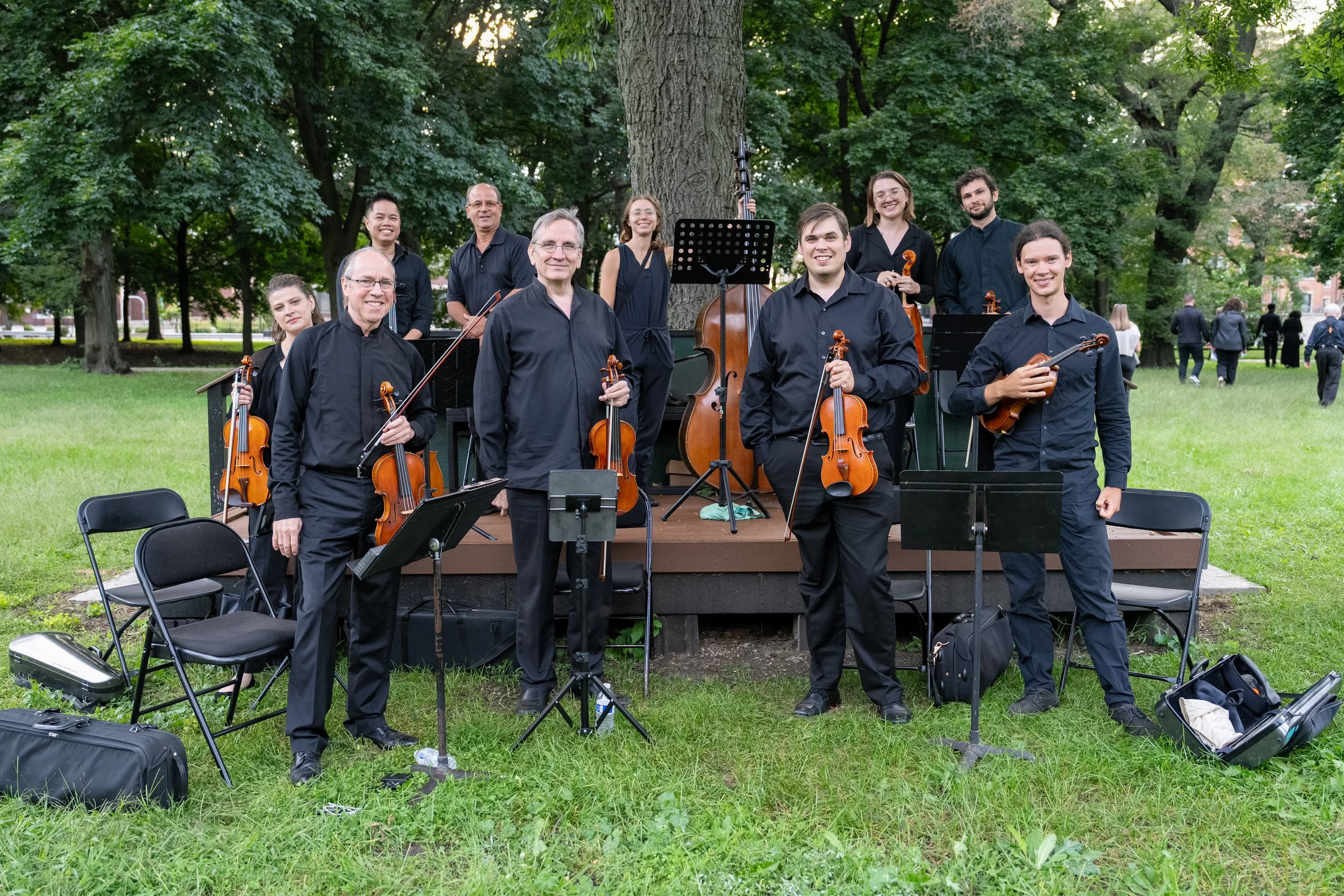 An orchestra of 10 musicians posing outdoors in a park with trees, holding string instruments such as violins, violas, and cellos.