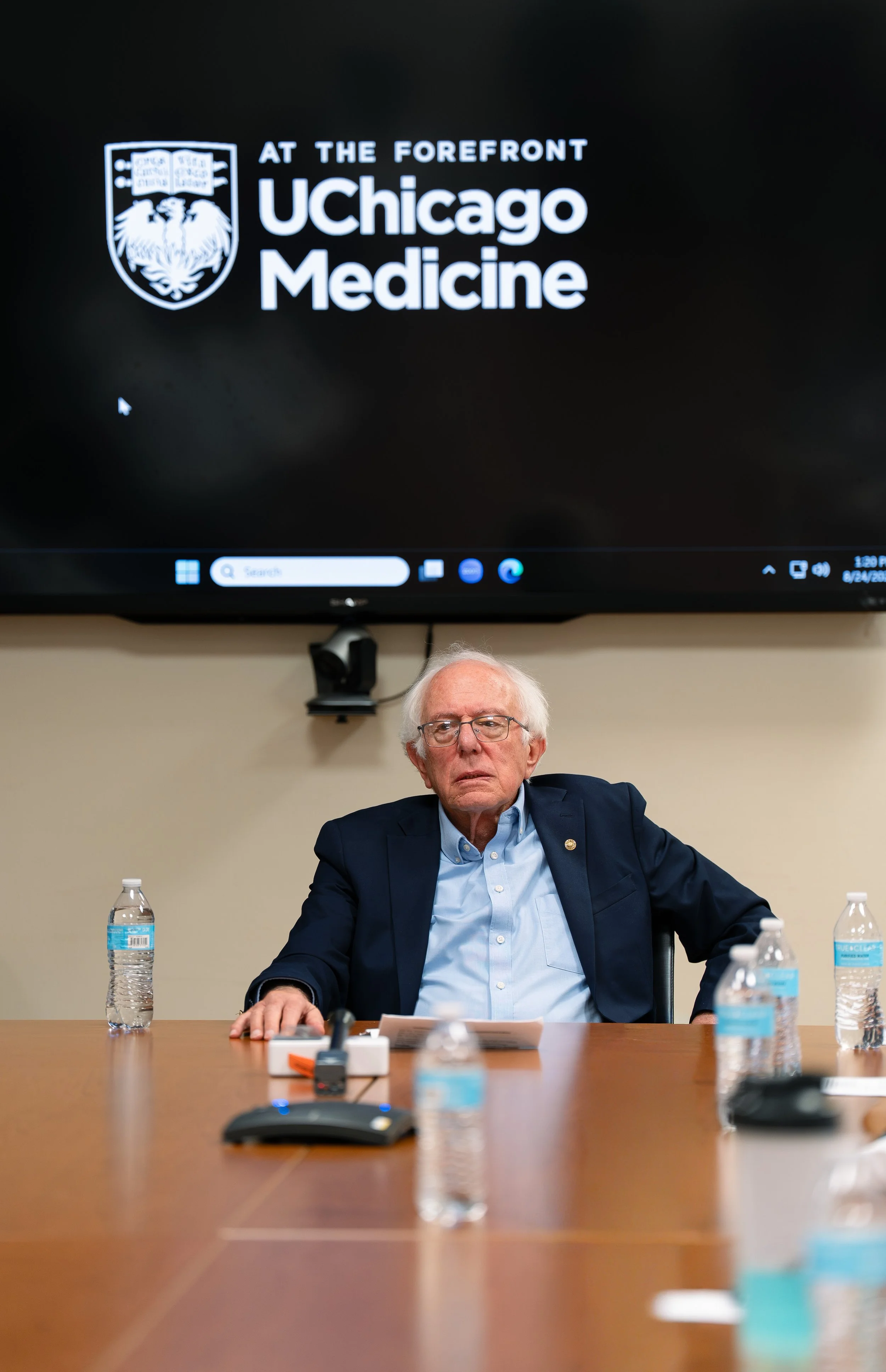 An elderly man with white hair and glasses sitting at a conference table with bottled water, a microphone, and papers, during a briefing at UChicago Medicine. A large screen behind him displays the UChicago Medicine logo.