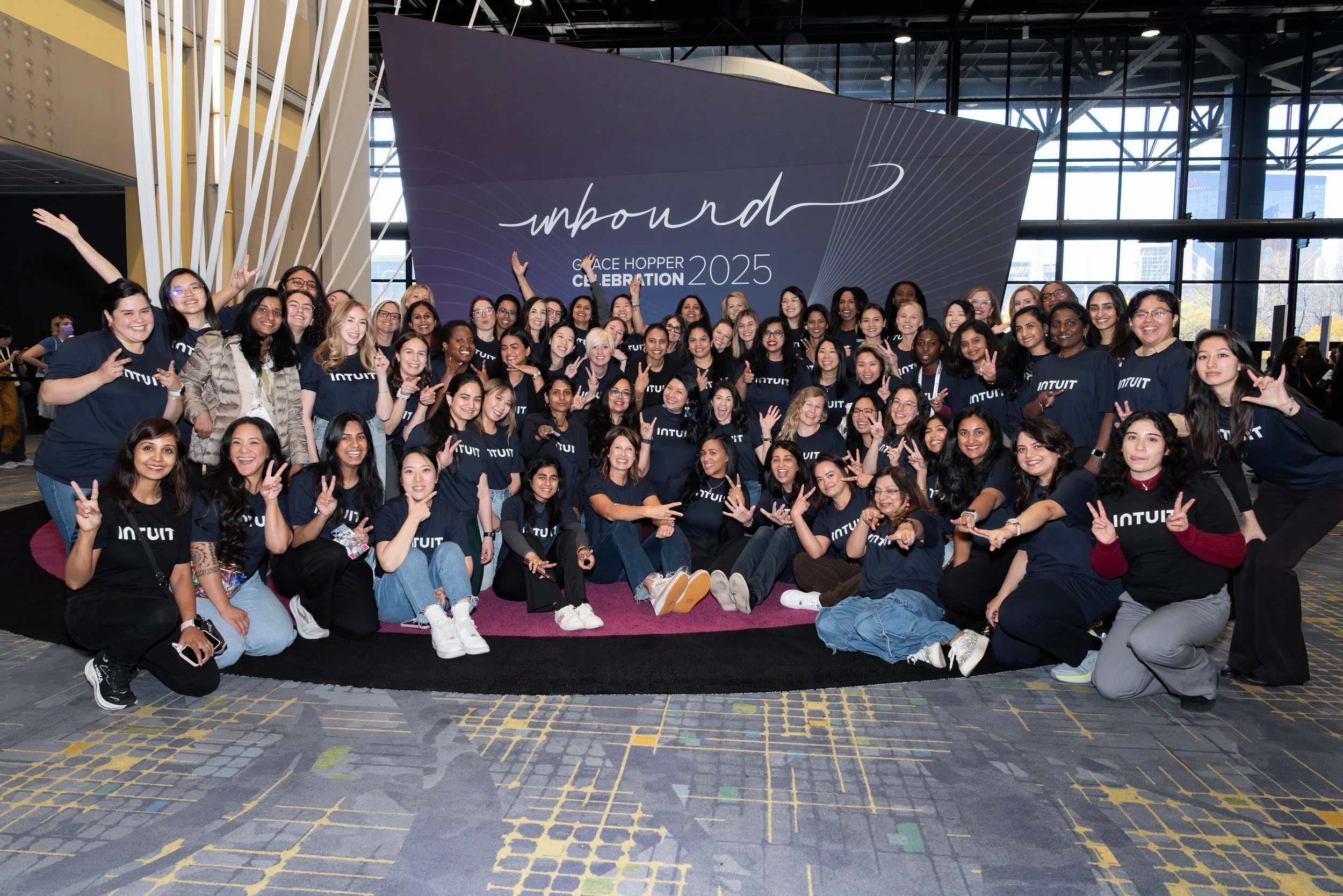 Group of women wearing matching navy blue T-shirts with 'INTUIT' logo, posing and smiling at a conference or event, with a large sign behind them that reads 'unbound Grace Hopper Celebration 2025'.