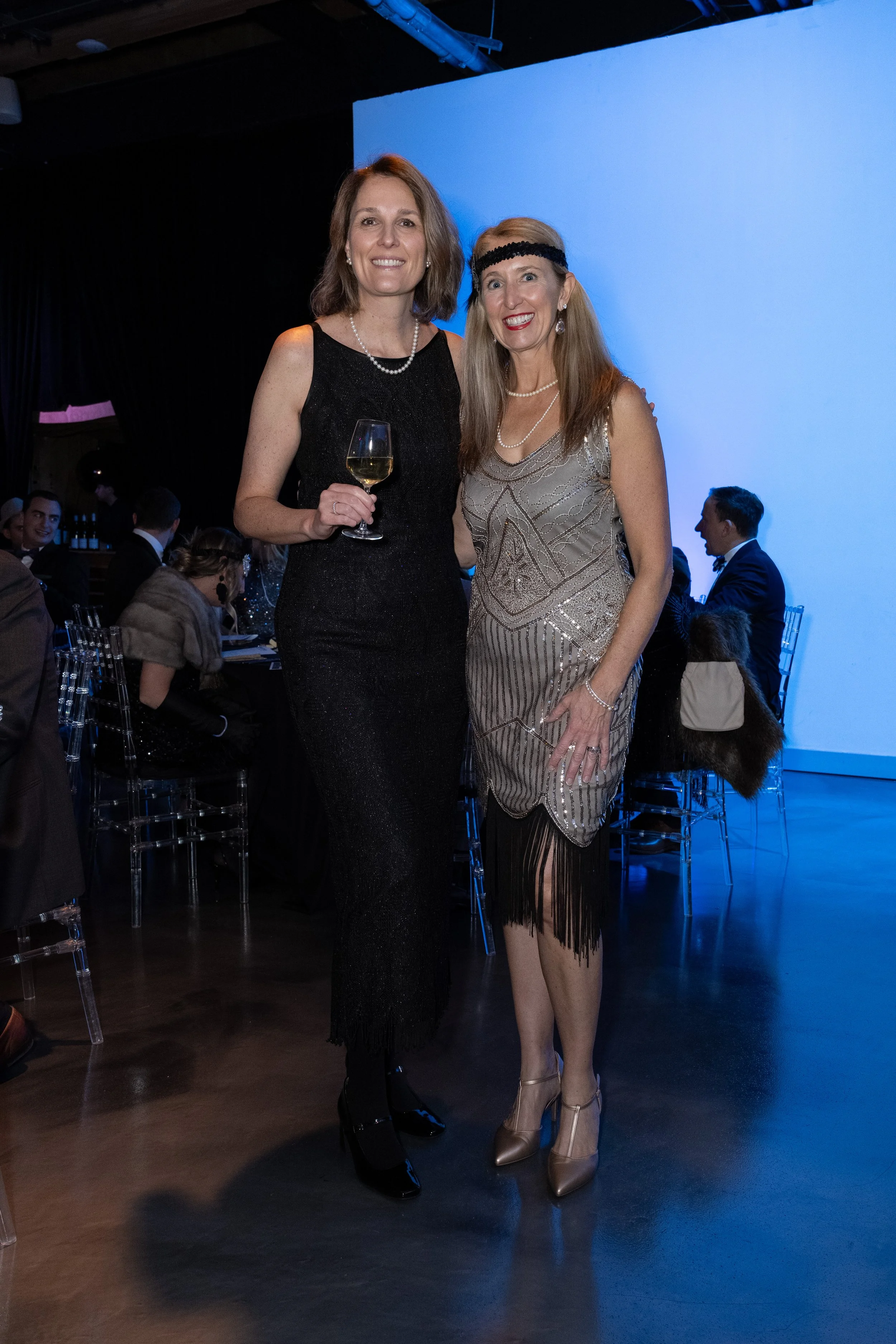 Two women dressed in elegant evening wear standing together at a formal event. One is holding a glass of white wine, and both are smiling at the camera.