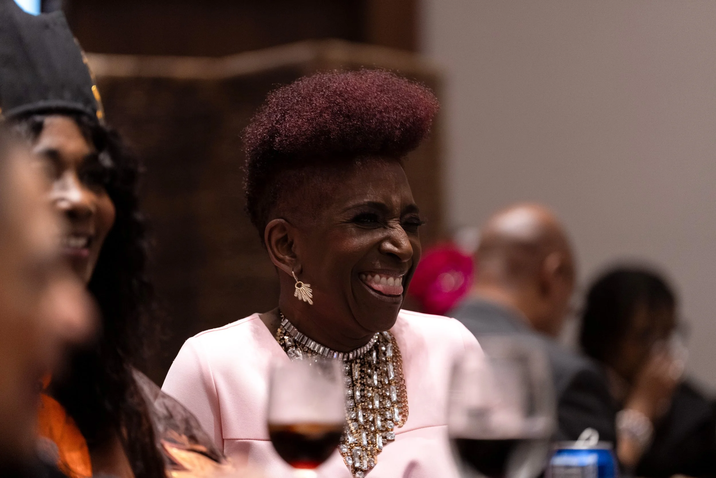 Woman smiling and laughing at a formal event, wearing a pink dress, jewelry, and earrings, with a glass of red wine in front of her.