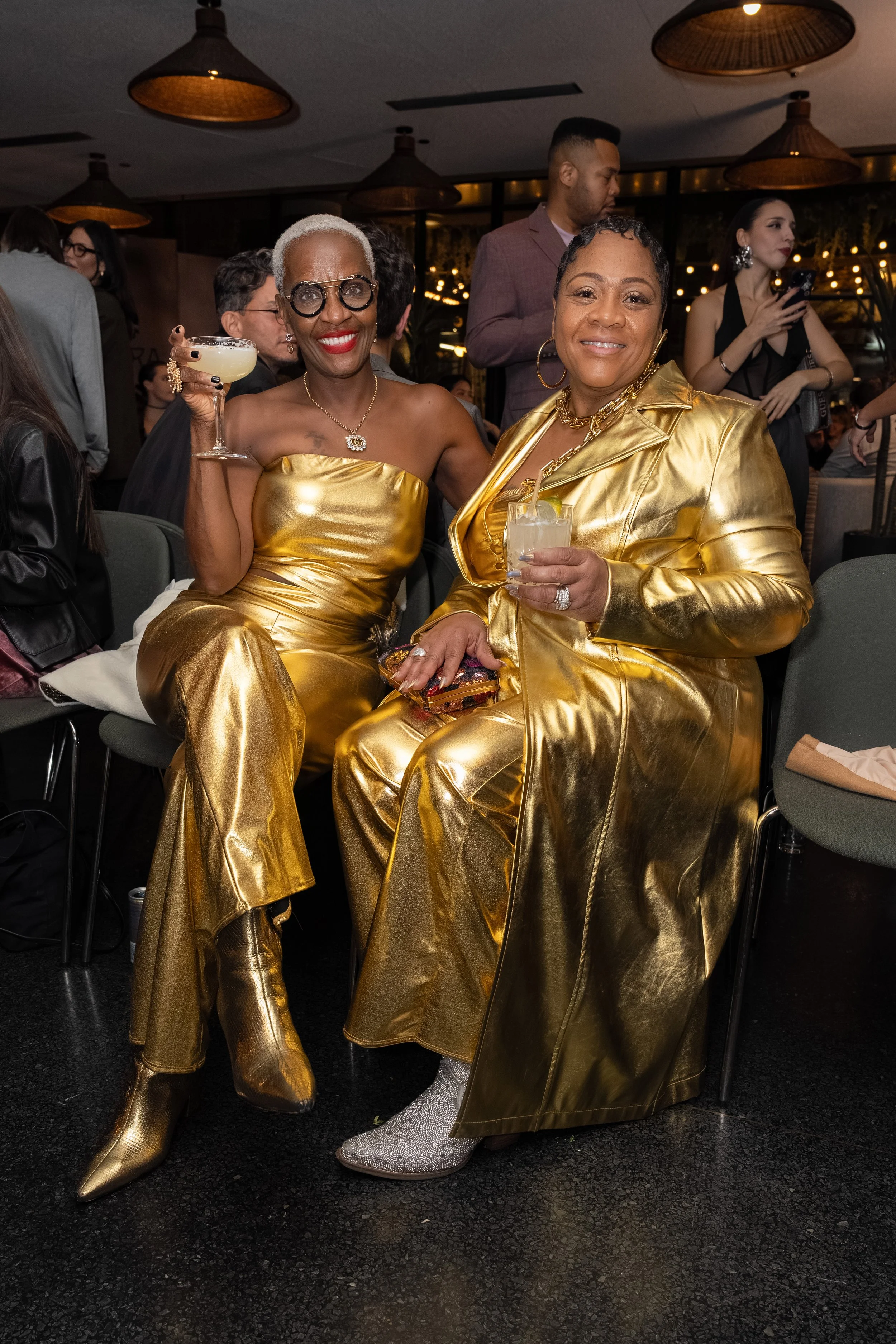 Two women dressed in shiny gold outfits sitting together at a party, holding drinks. The background shows other guests, dim lighting, and decorative hanging lights.