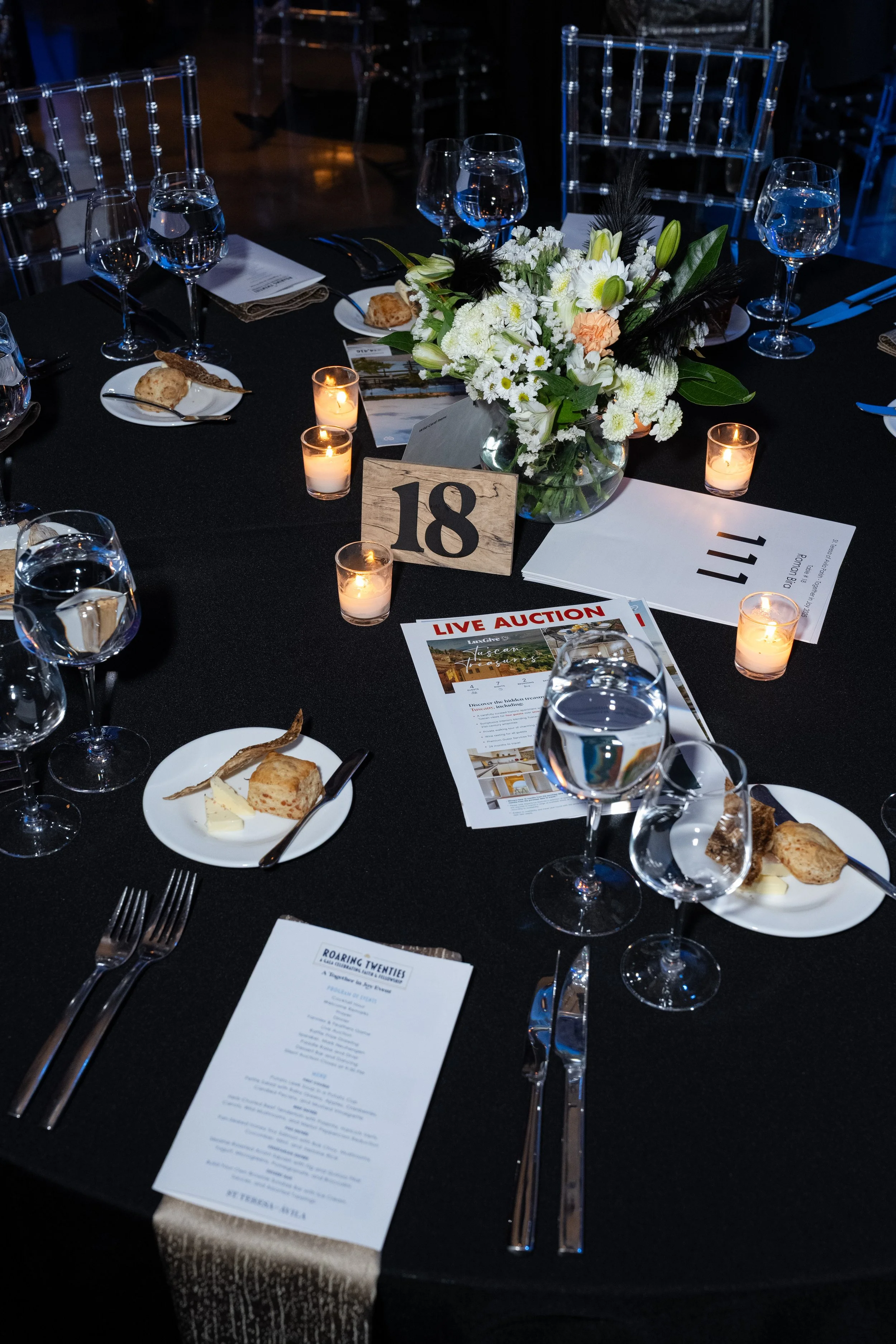 A formal event table setting with black tablecloth, white floral centerpiece, lit candles, plates of bread and cheese, wine glasses filled with water, printed event programs, and a table number card with the number 18.