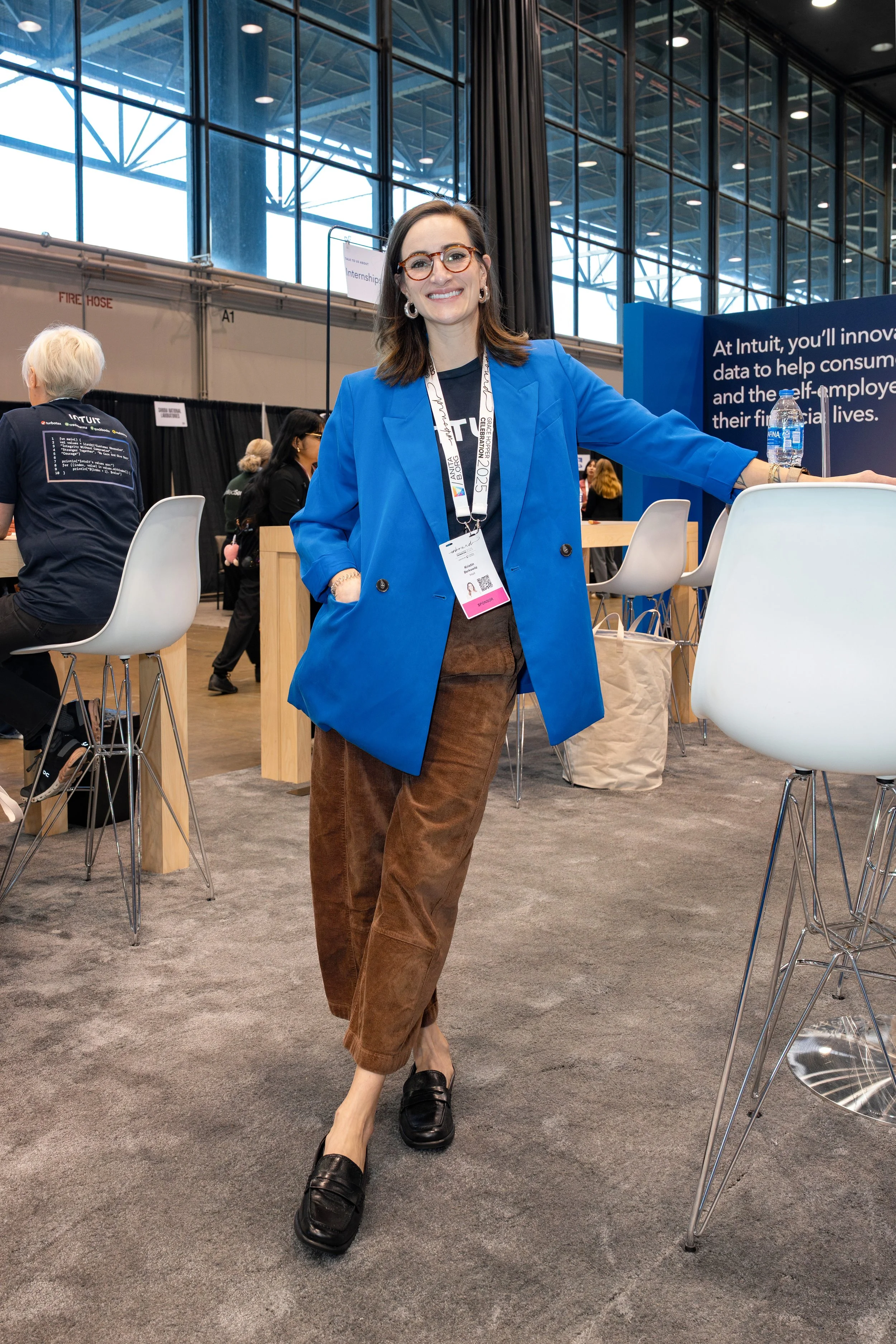 A woman wearing glasses, a blue blazer, and brown pants standing at a conference booth with a smile.
