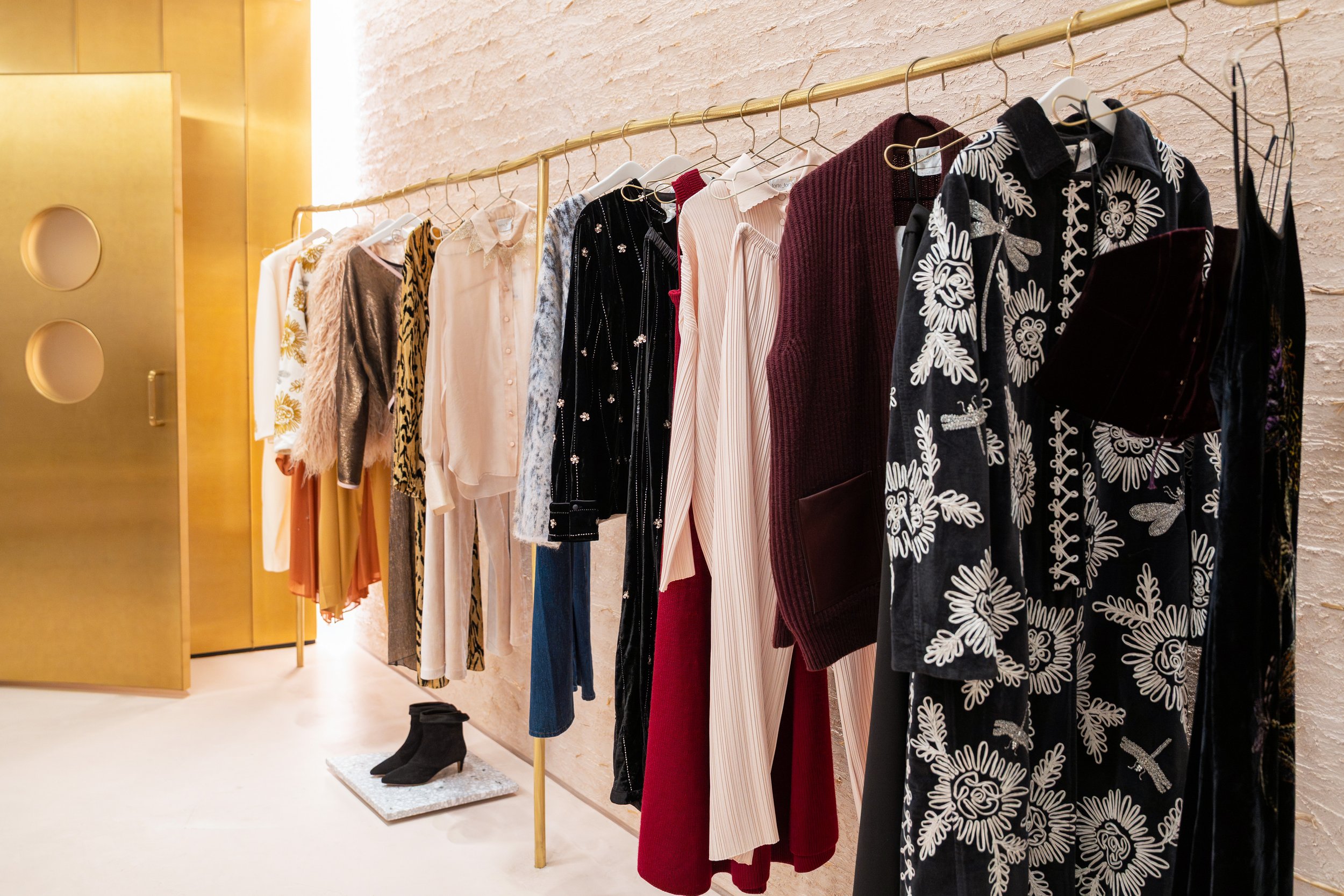 Clothes hanging on a gold clothing rack in a boutique, with various fashionable dresses, blouses, and outerwear, and a pair of black ankle boots on the floor.