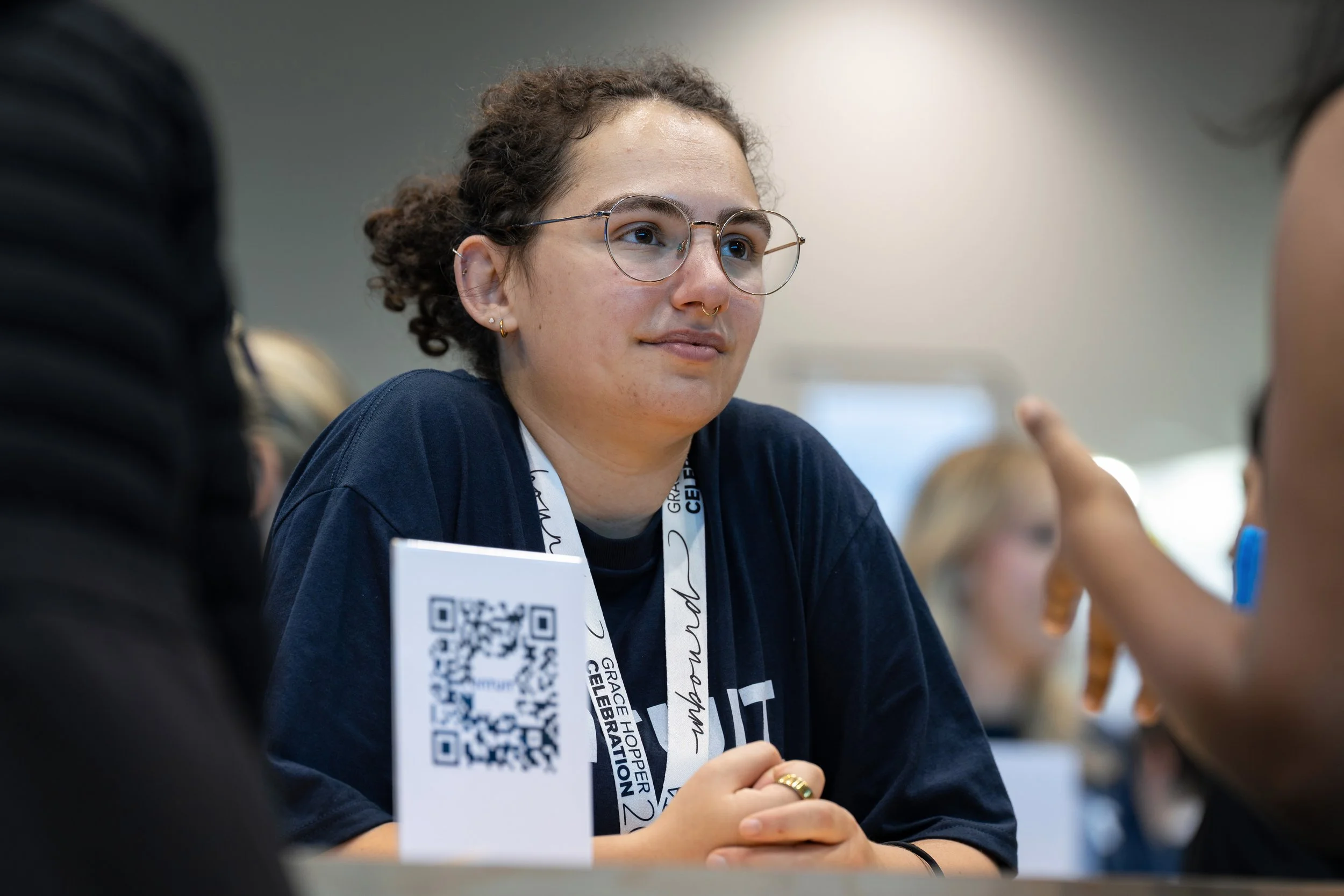 A young woman with glasses and curly hair sitting at a table during an event. She is wearing a navy shirt and has a lanyard with event branding. Others are engaged in conversation around her.