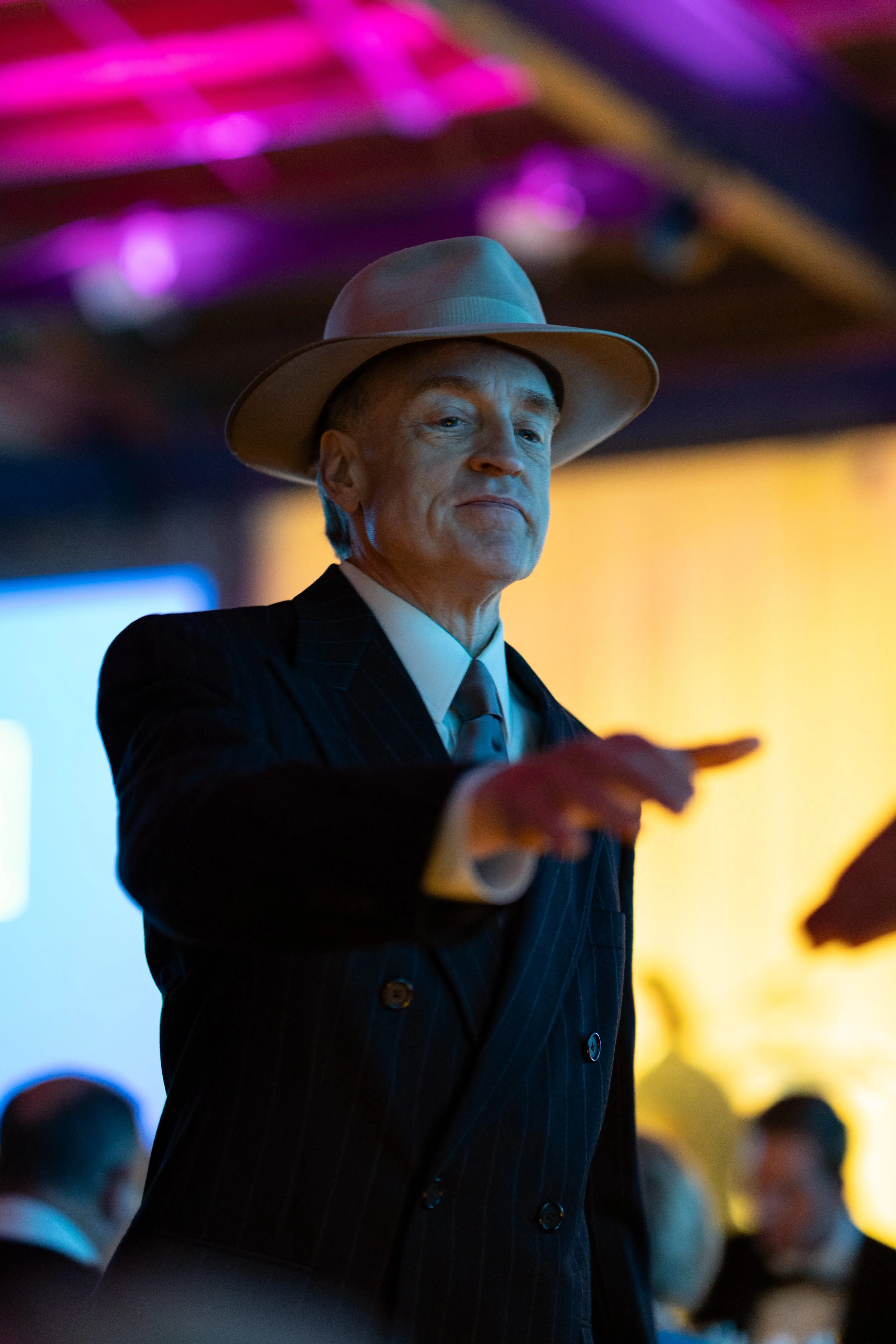 An older man in a dark pinstripe suit and a light-colored fedora pointing with his right hand at an event with colorful background lights.