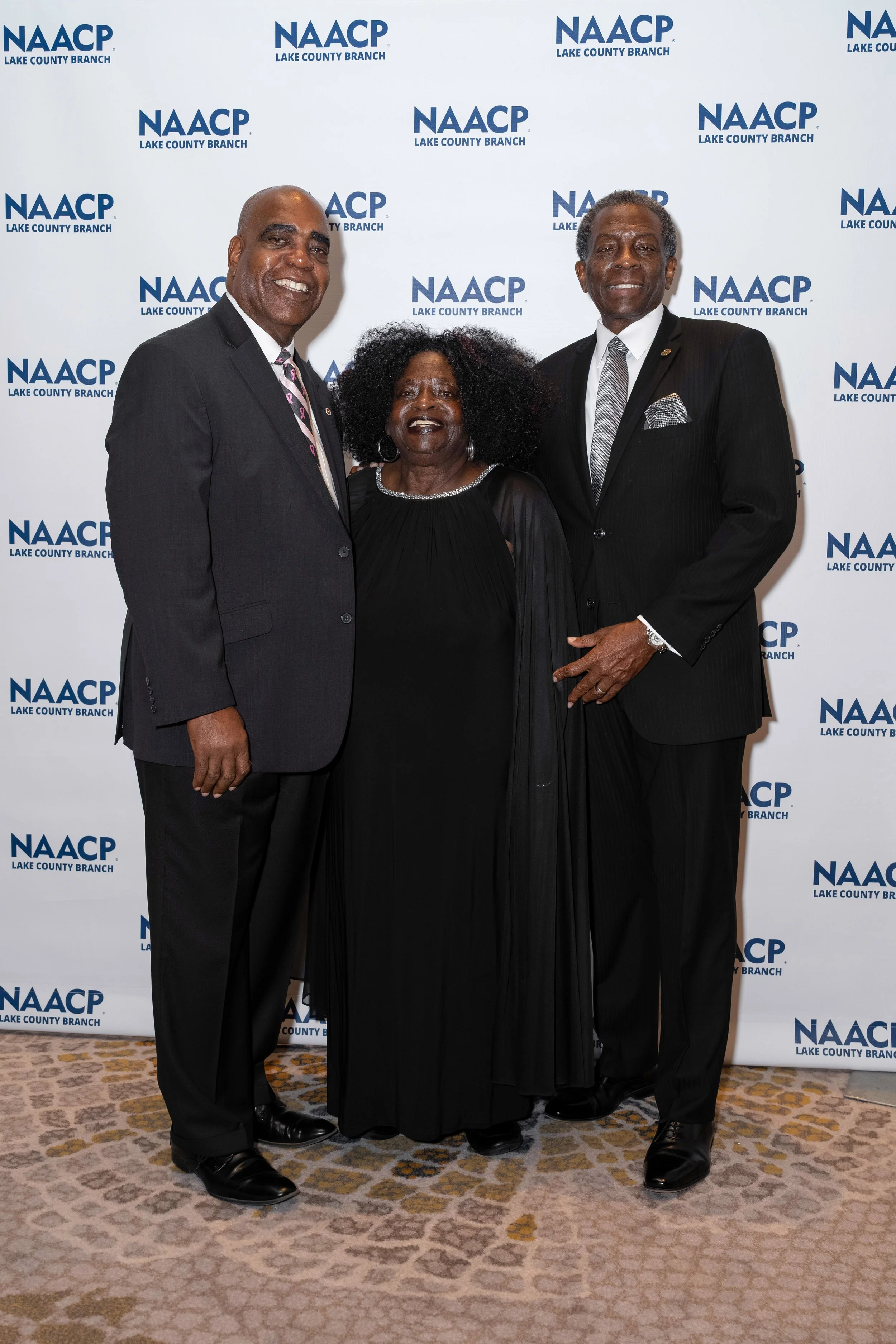 Three individuals dressed in formal attire, standing together and smiling at an event, with a backdrop displaying the NAACP Lake County Branch logo repeated multiple times.
