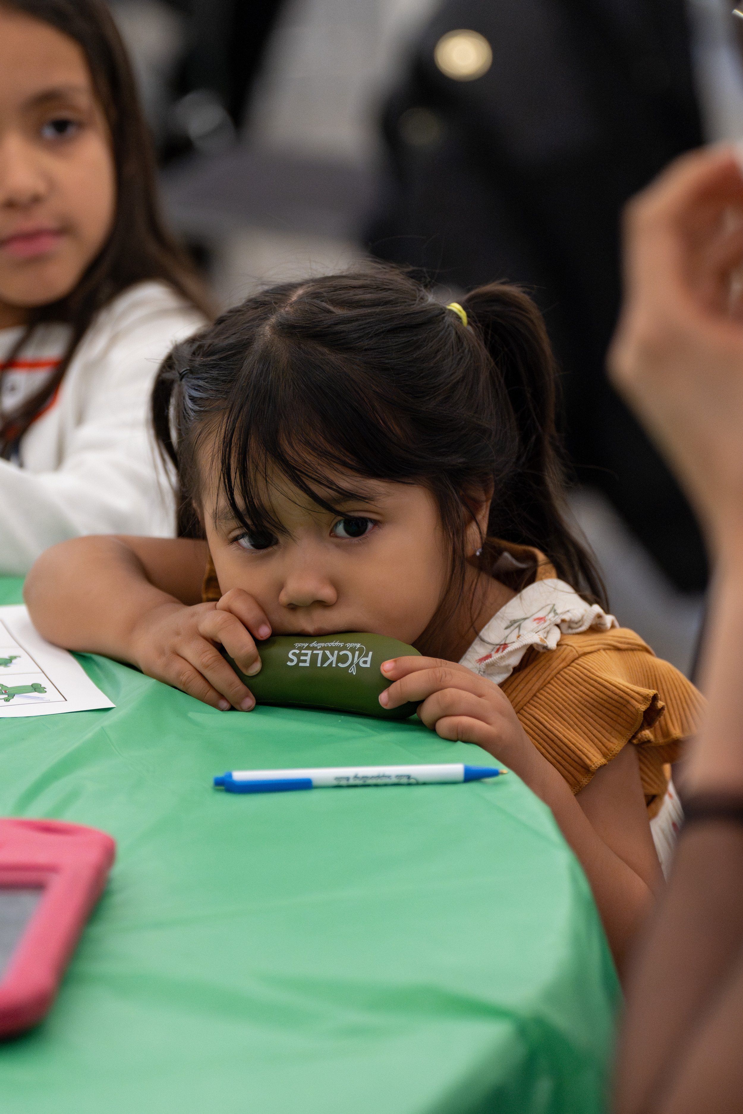 Young girl with dark hair in a ponytail, wearing a brown shirt with ruffles on the sleeves, resting her head on her arms on a green table, holding a green pepper-shaped object labeled 'Pickles,' looking directly at the camera. Other children and a pe