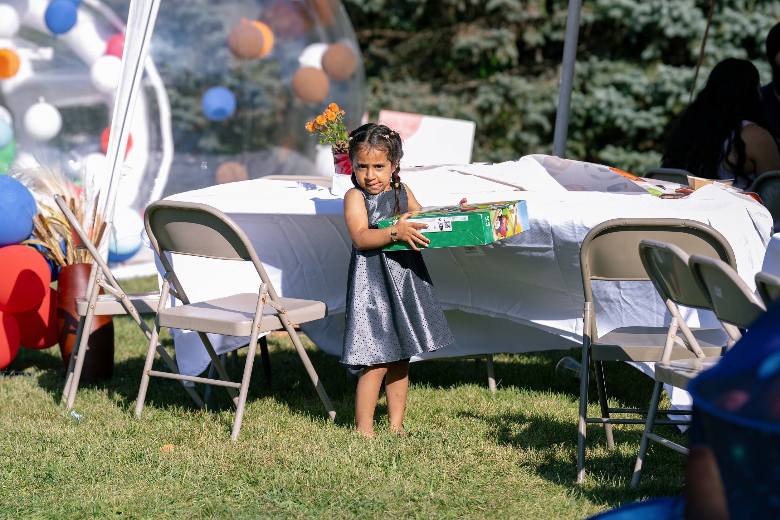 A young girl in a black dress holding a board game at an outdoor celebration with decorations and a table covered with a white cloth.