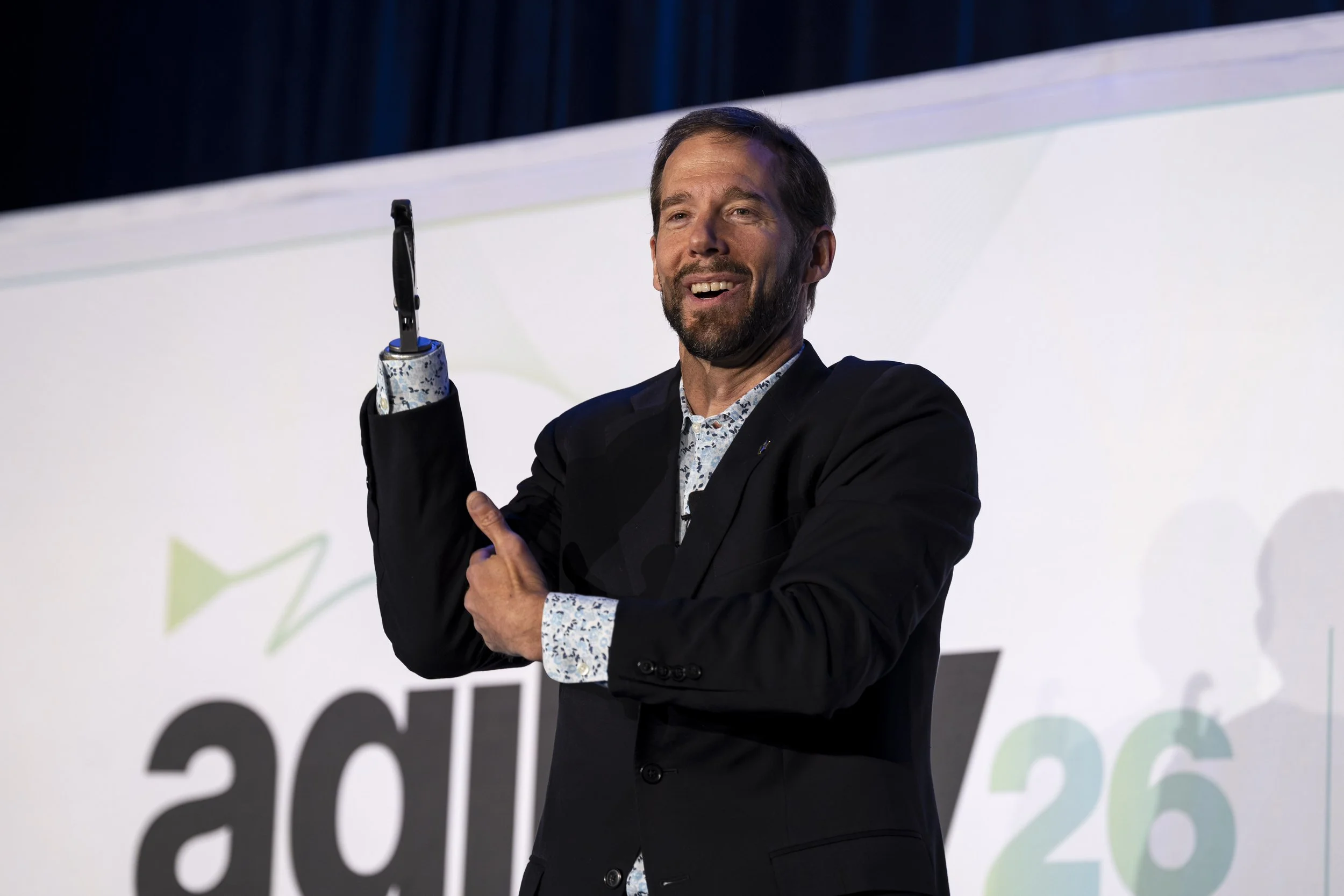 A man with a beard and dark hair, smiling, wearing a dark suit and a patterned shirt, is on stage with a microphone in his right hand, speaking or presenting.
