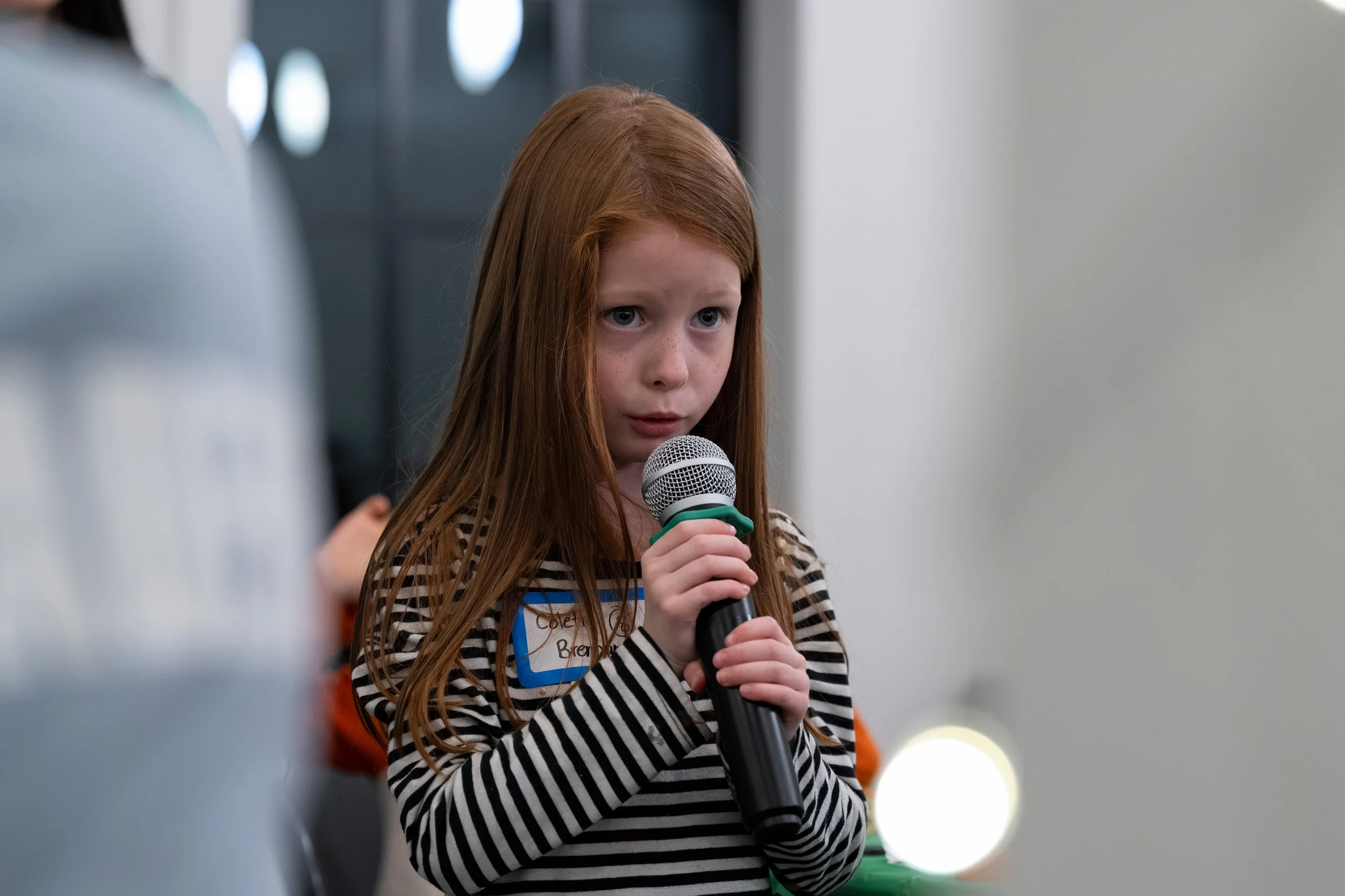 A young girl with long red hair and freckles is holding a microphone and speaking at an indoor event, wearing a black and white striped shirt.