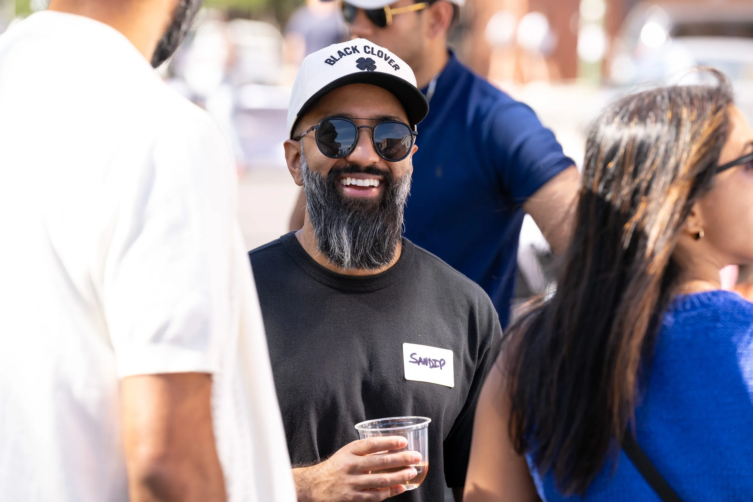 A man with a beard, wearing sunglasses, a white cap that says 'Black Clover', a black t-shirt with a name tag that reads 'Sandeep', and holding a clear plastic cup, is smiling at an outdoor social gathering.
