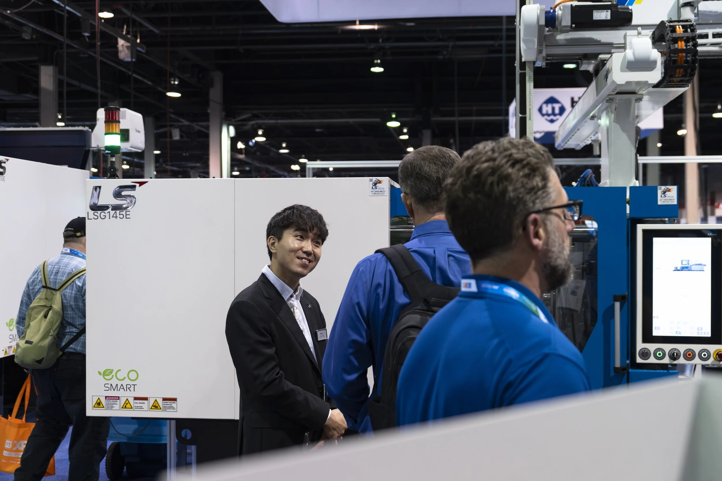 People at an exhibition booth engaging with a machine labeled 'LSG145E' and 'eco SMART', with a woman in a black suit smiling and a man in blue in front of a monitor, in an indoor convention center.