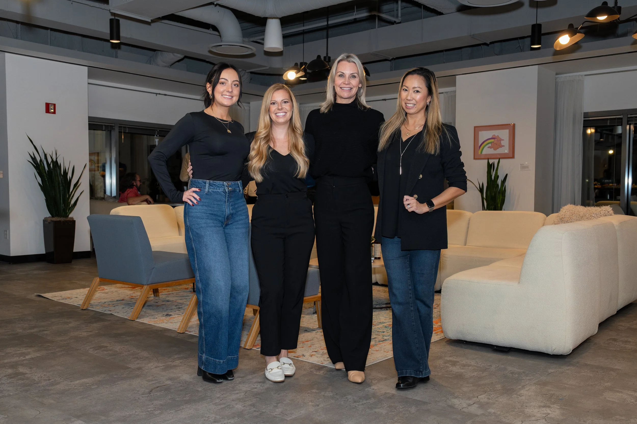 Four women standing together in a modern, well-lit indoor lounge area, smiling at the camera. They are dressed in black tops and jeans or black pants, with plants and artwork visible in the background.