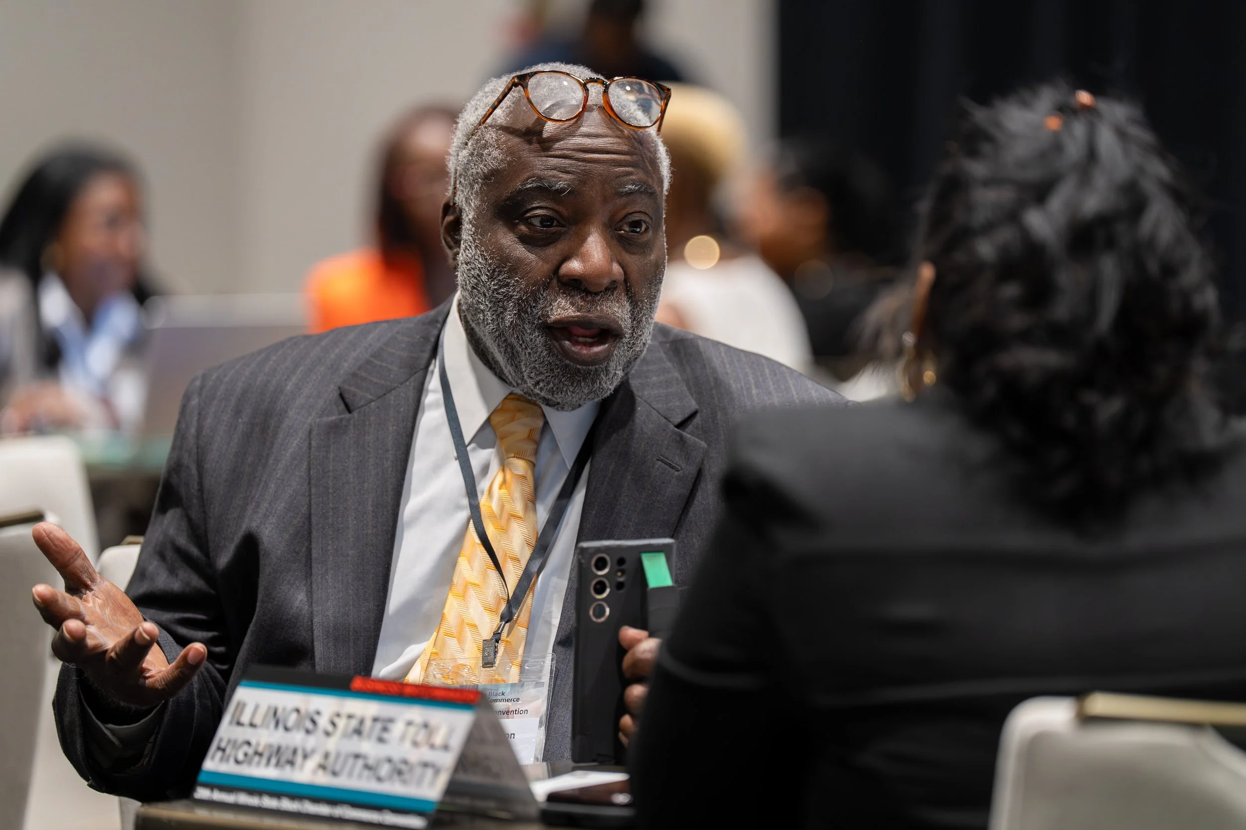An elderly man with gray hair, beard, and glasses on his head, wearing a dark suit and yellow tie, talking to a woman with dark hair in an indoor conference setting. There are other people blurred in the background.