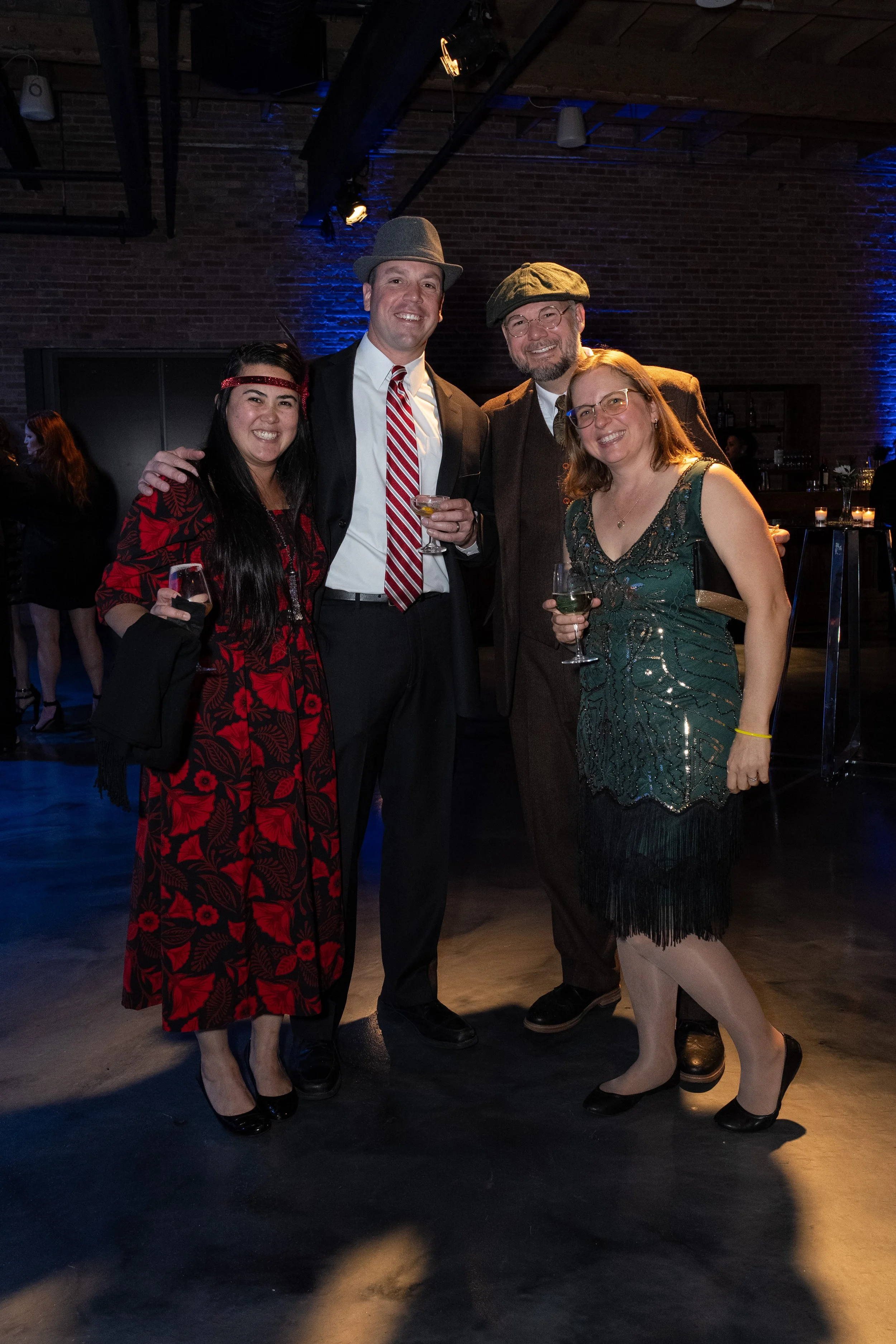 Four people dressed in vintage or themed attire posing with drinks at an indoor event with dark lighting and brick walls.