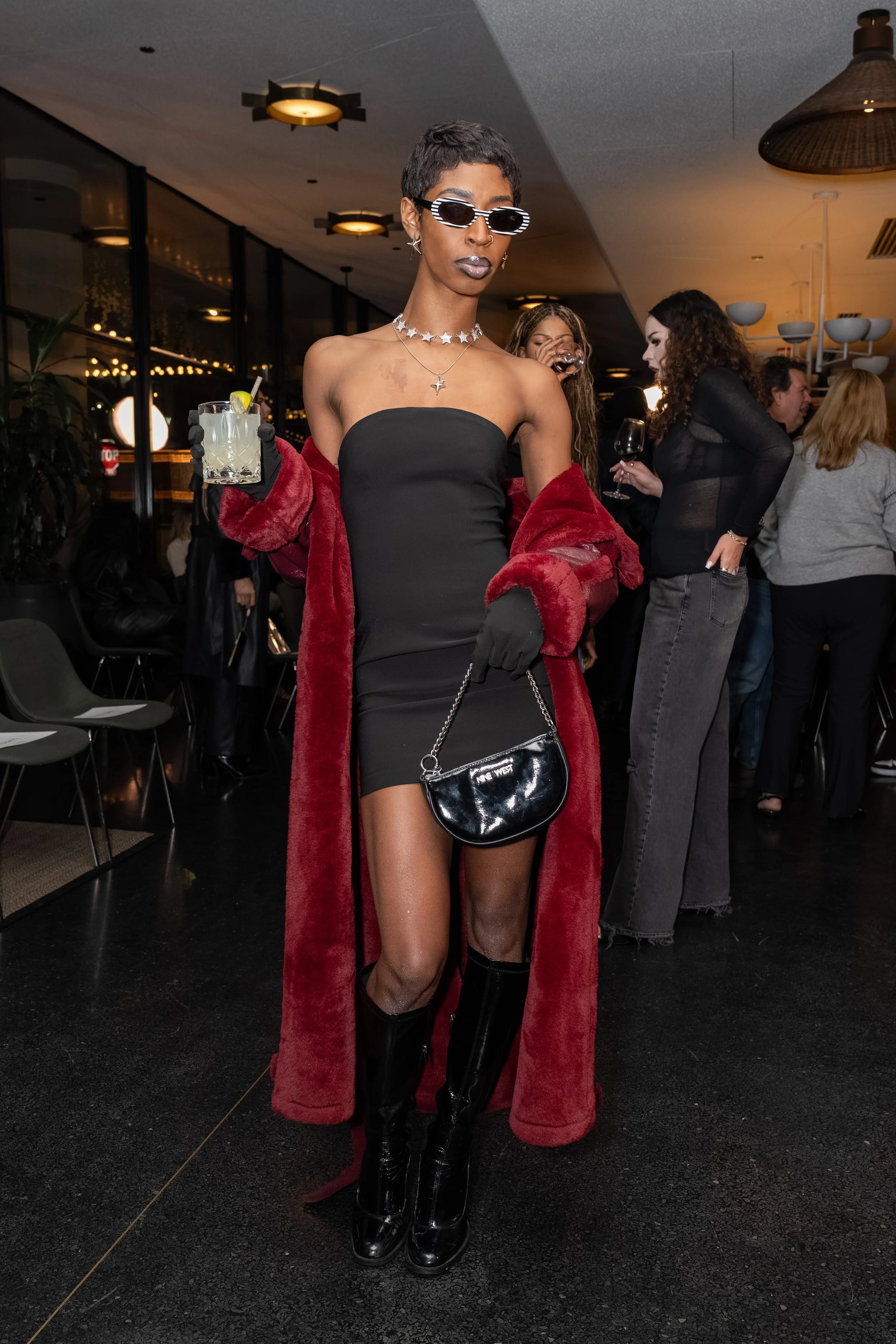 A fashionable woman at a social event wears a strapless black dress, thigh-high black boots, large sunglasses, layered necklaces, and a red coat draped over her shoulders. She holds a cocktail in one hand and a black purse in the other, with a stylis