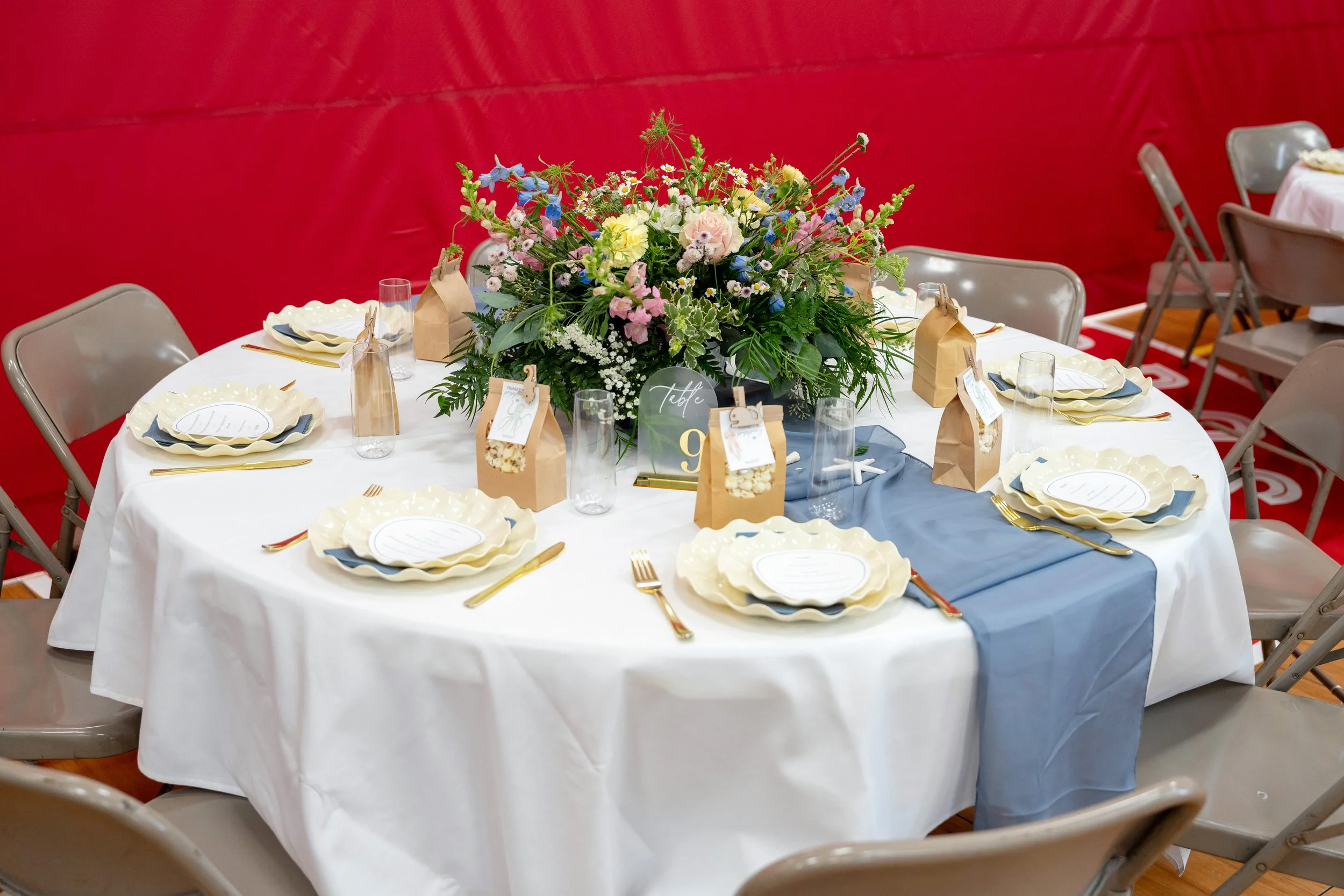 Round table set for a celebration with a large floral centerpiece, six place settings with cream-colored plates, gold-colored utensils, clear glasses, and brown paper gift bags, with a blue table runner, against a red background.