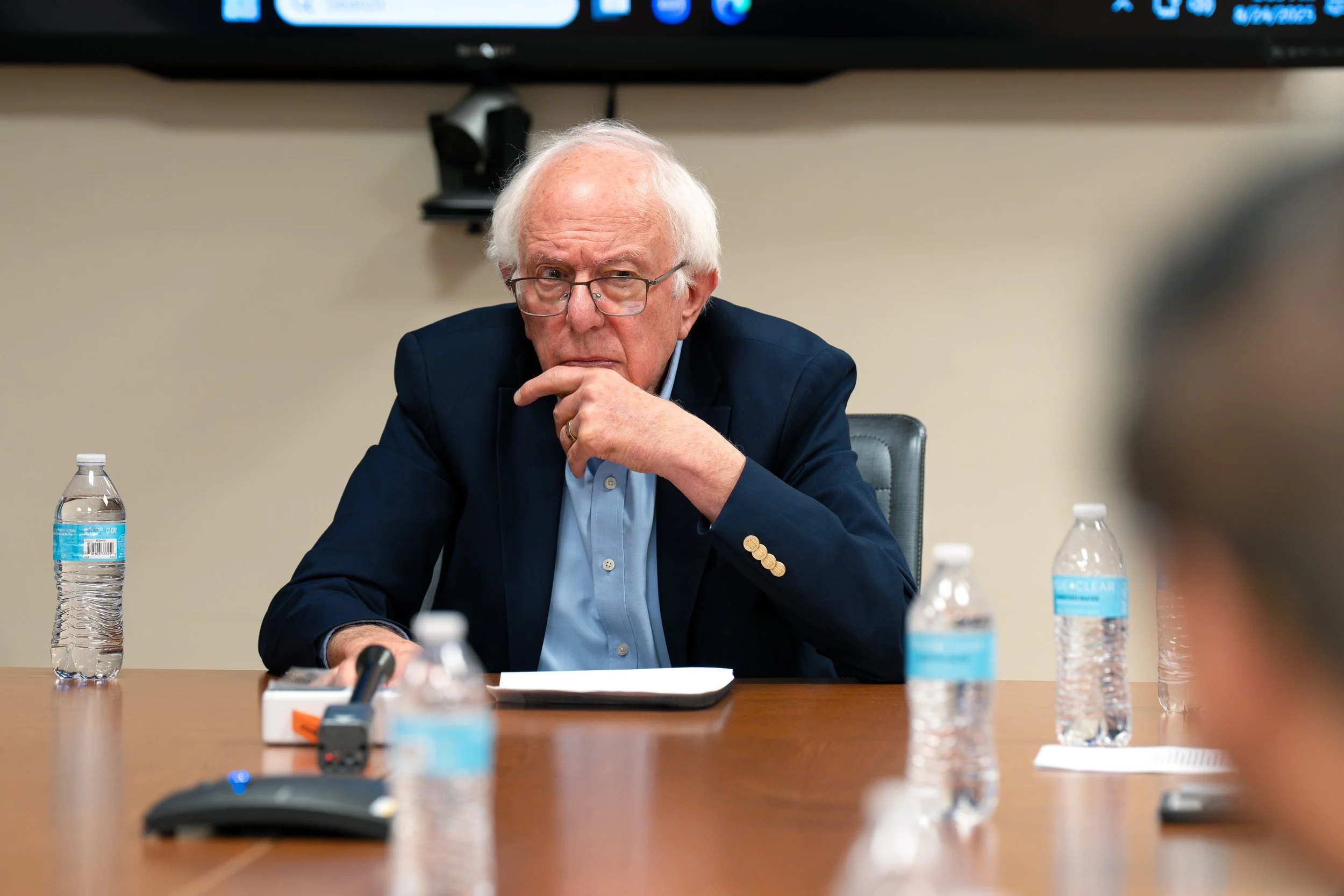 An elderly man with white hair and glasses, wearing a navy blazer and light blue shirt, sits at a conference table with his hand on his chin, appearing thoughtful. There are water bottles, a microphone, and papers on the table. The background shows a
