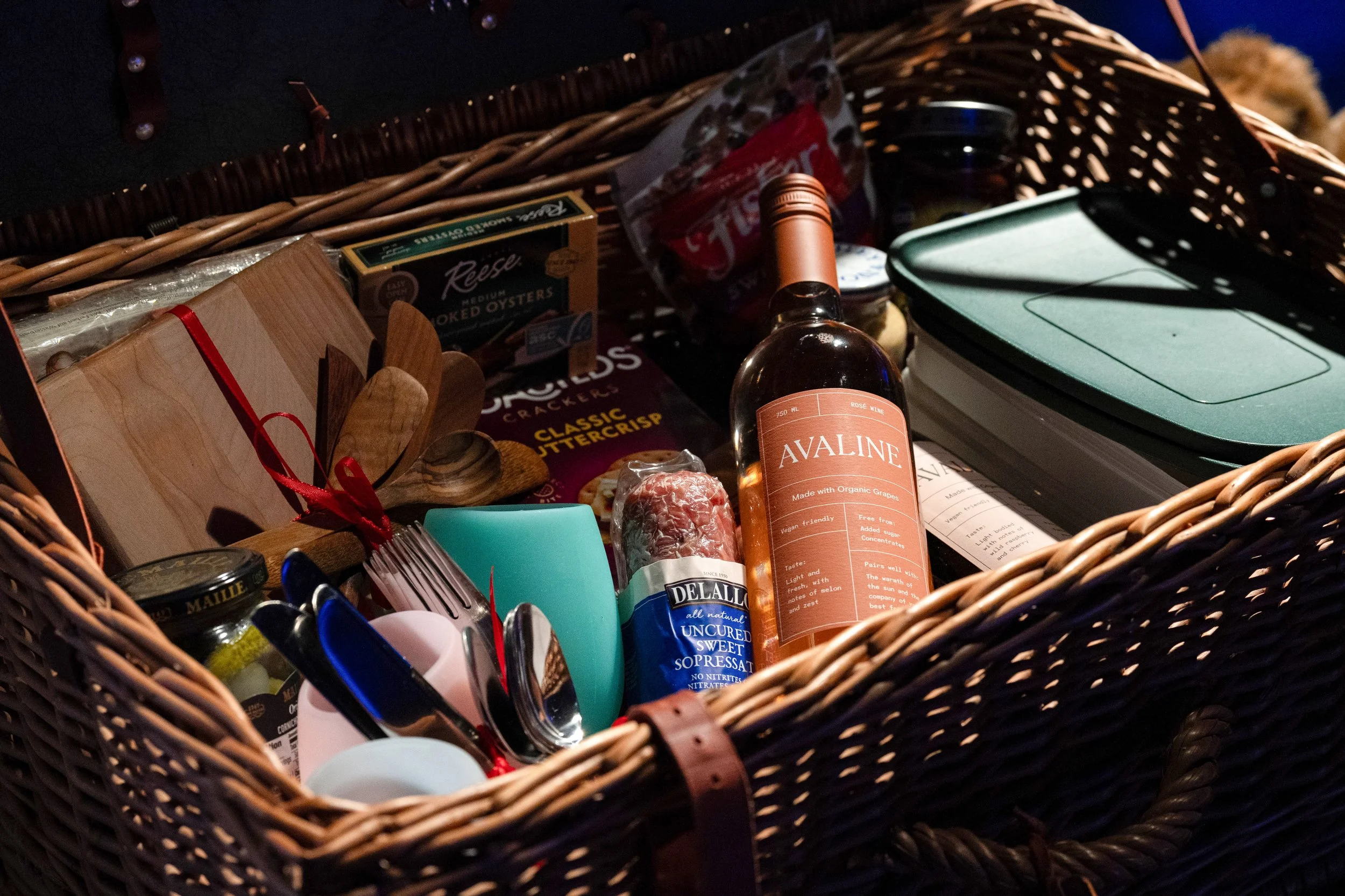 A wicker basket filled with assorted food items and utensils, including a bottle of Avaline wine, a package of uncured sweet sopressata, a box of Reese smoked oysters, a wooden cutting board, a set of knives, and other packaged snacks and supplies.