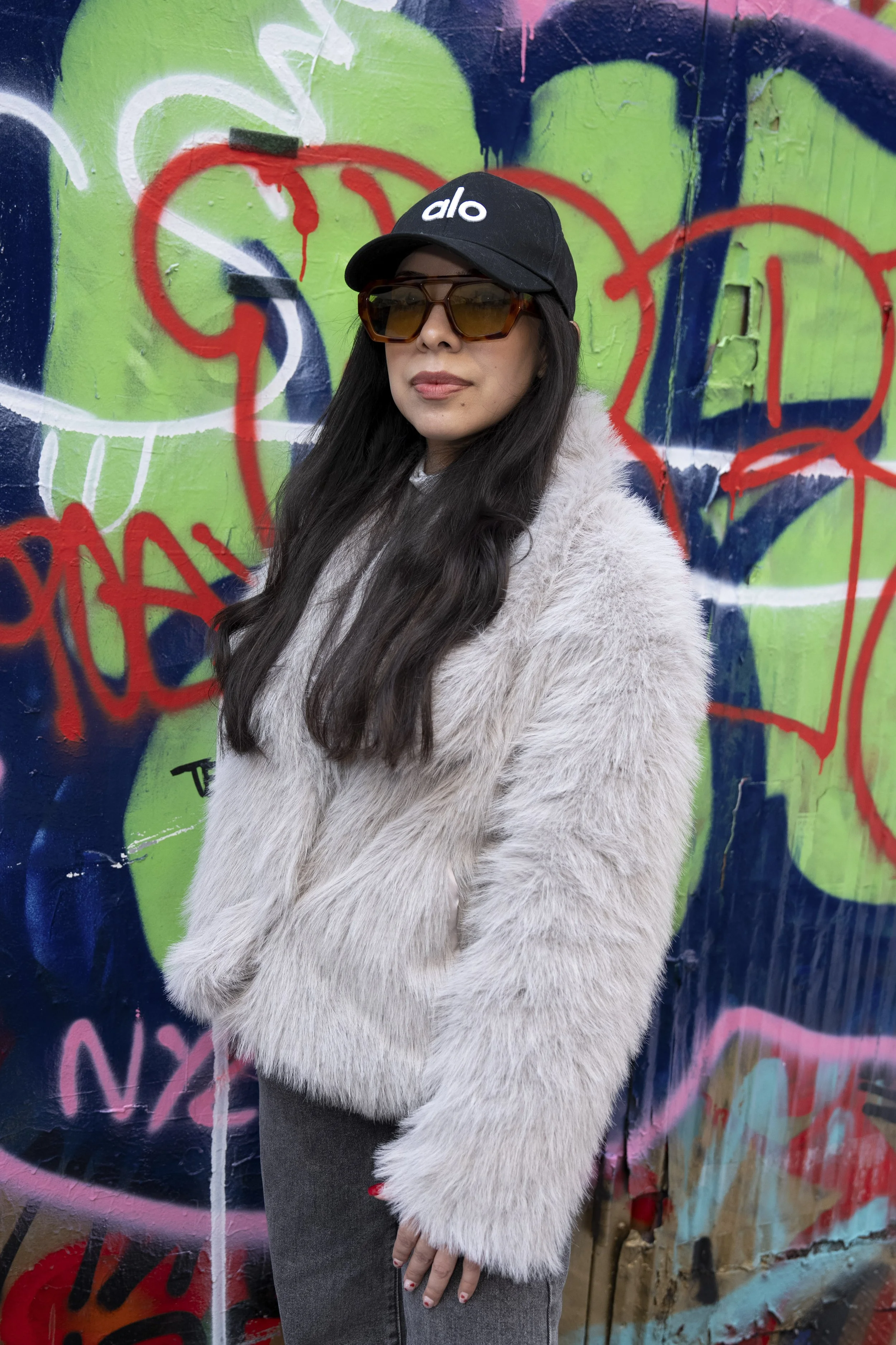 A woman with long dark hair wearing a black cap with white lettering, sunglasses, a fluffy light-colored coat, and gray pants standing in front of a colorful graffiti wall.