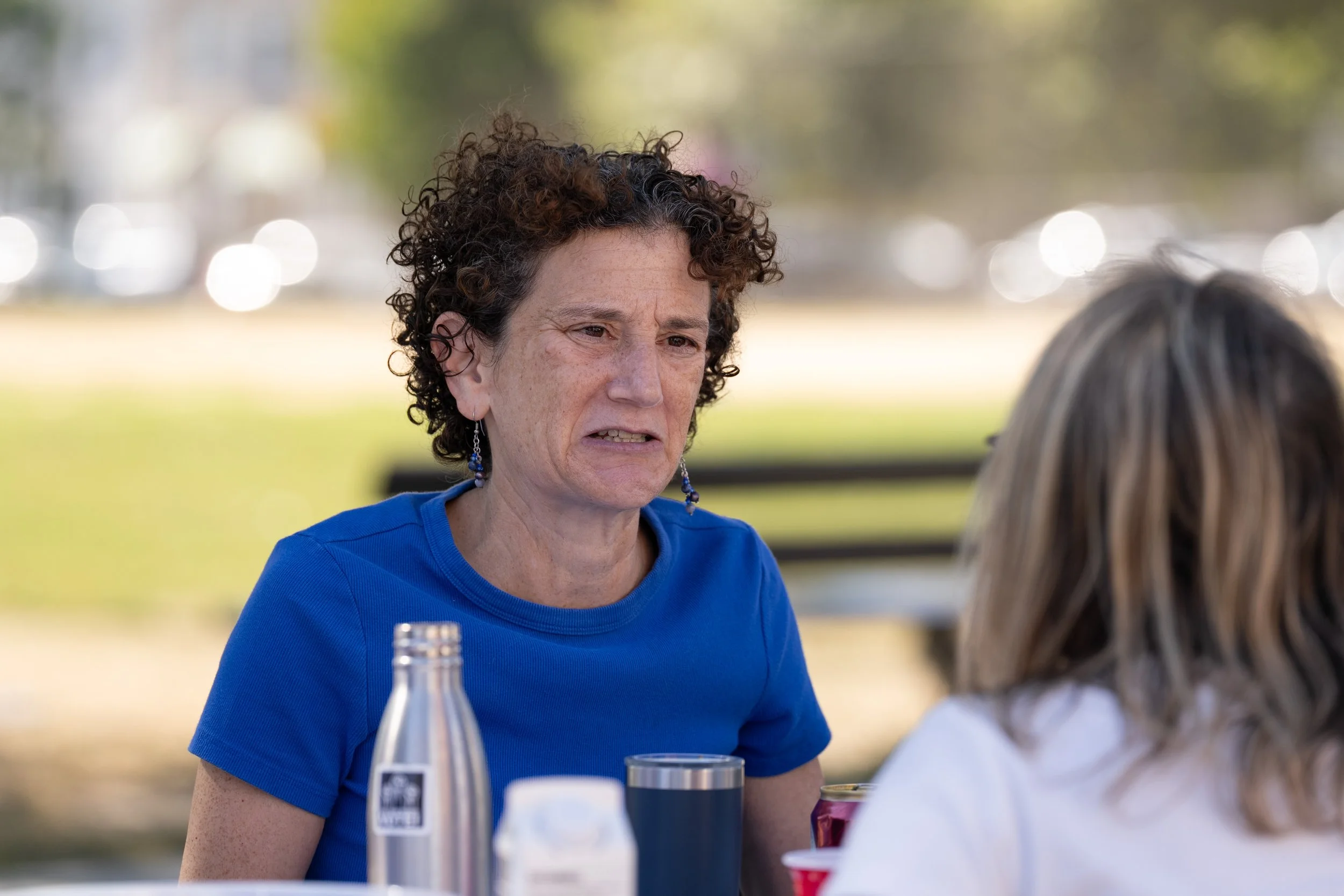 A woman with curly hair wearing a blue shirt and earrings is sitting at a table outdoors, speaking to another woman with blonde hair, with a park and trees in the background.