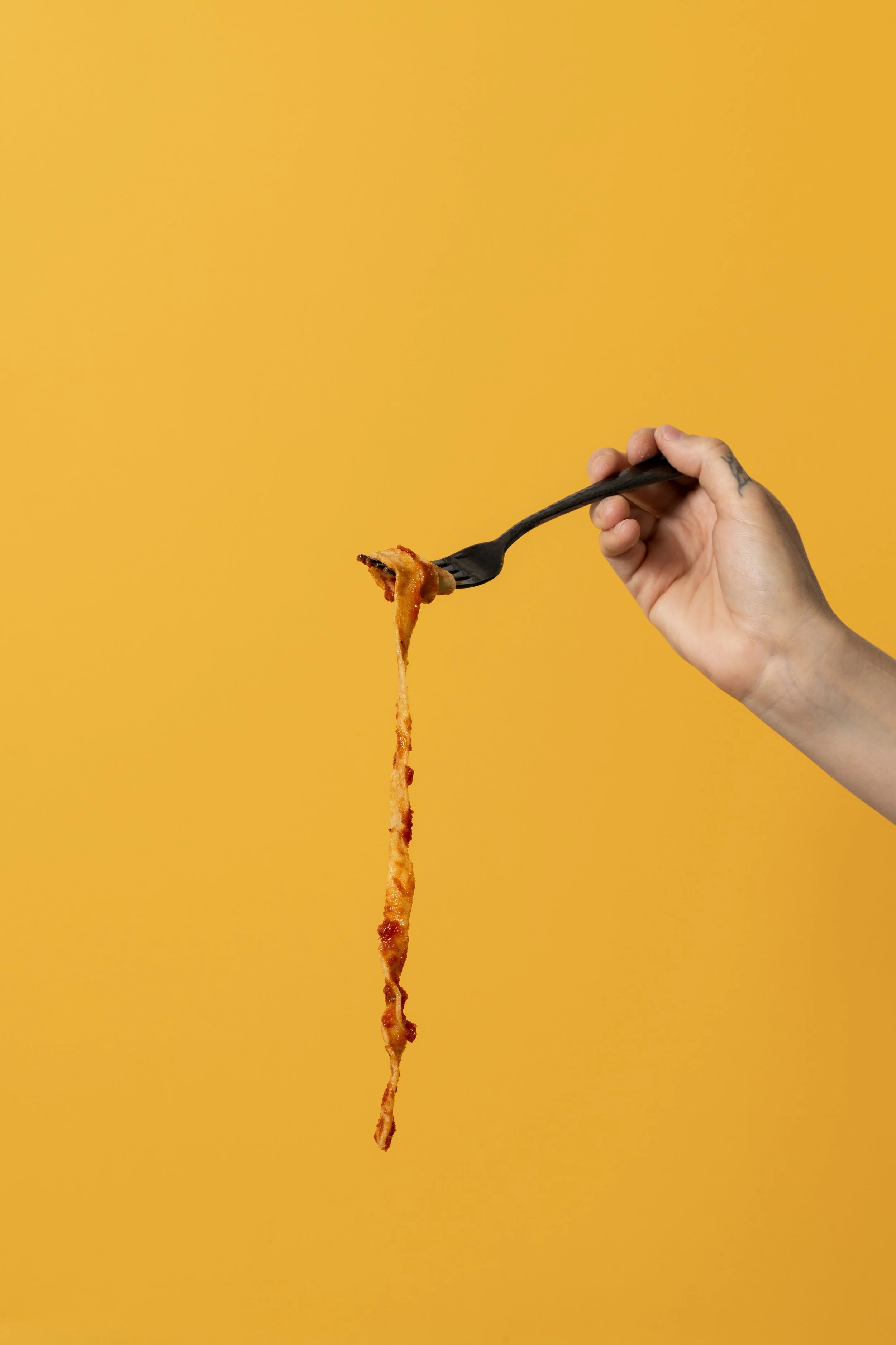 A hand with a tattoo on the wrist holding a black fork with a strand of melting cheese and pepperoni, against an orange background.