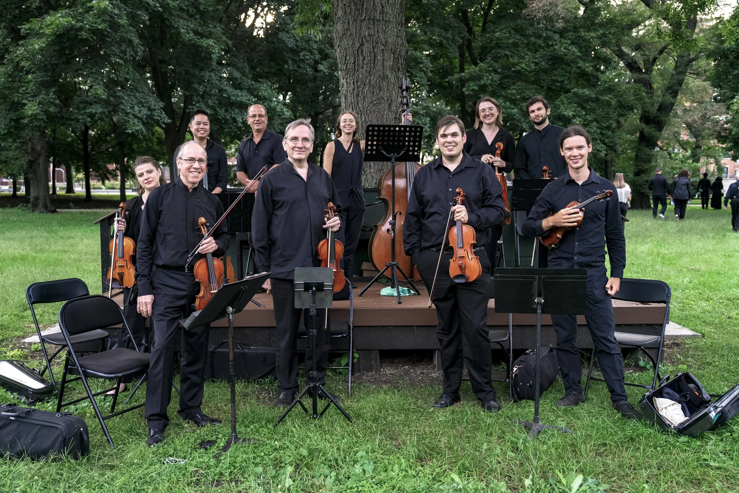 A group of musicians standing outdoors under a large tree, holding their string instruments, with music stands and chairs around them in a park setting.