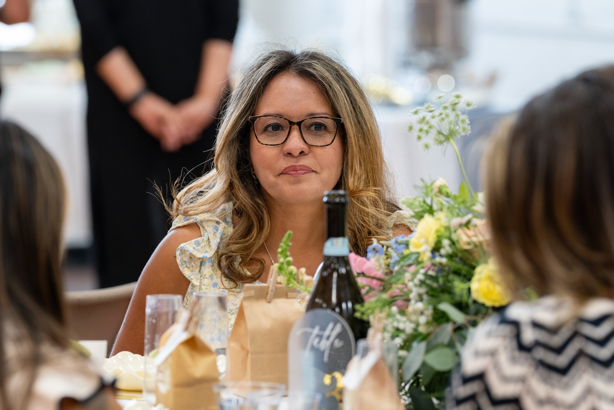 A woman with glasses and light brown wavy hair looking at the camera during a gathering or celebration, surrounded by flowers, drinks, and people at a table.