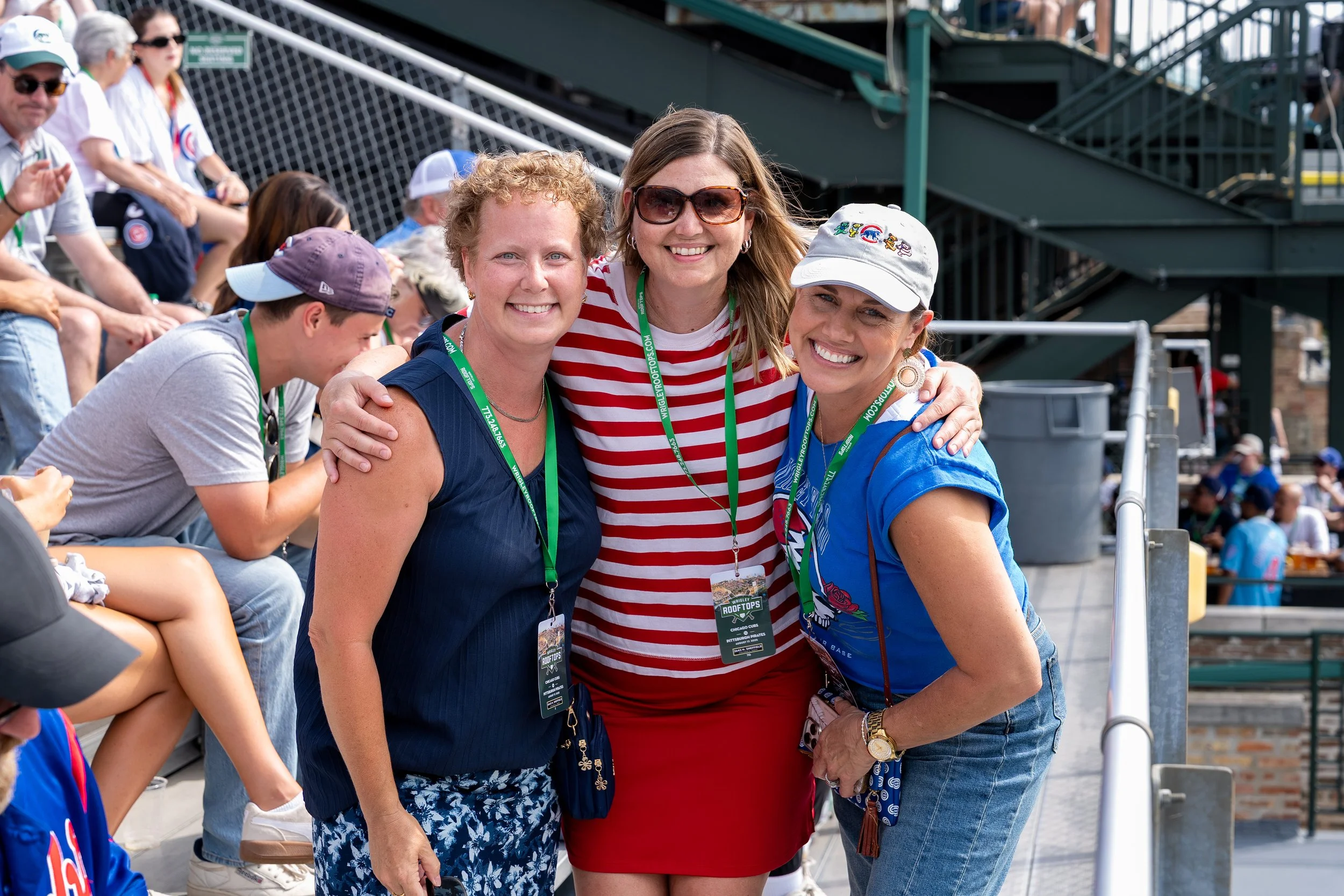 Three women smiling and posing together at an outdoor event, with people sitting in bleachers in the background. All three women are wearing event lanyards.